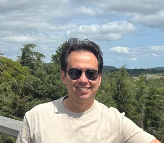 Richard Webeling, wearing sunglasses and a beige t-shirt smiles while standing outdoors in front of a background of trees.
