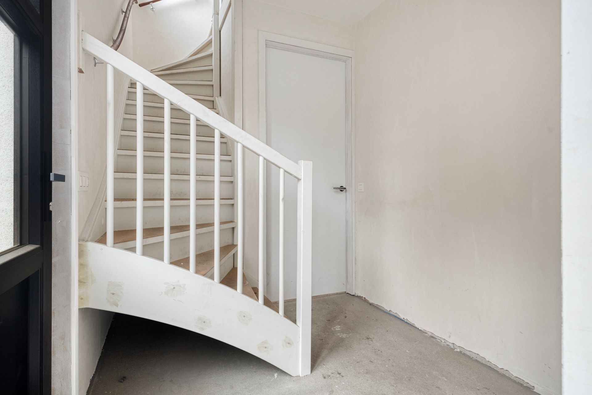 Een witte wenteltrap in een lege kamer leidt naar boven. Bovenaan is een gesloten deur zichtbaar.