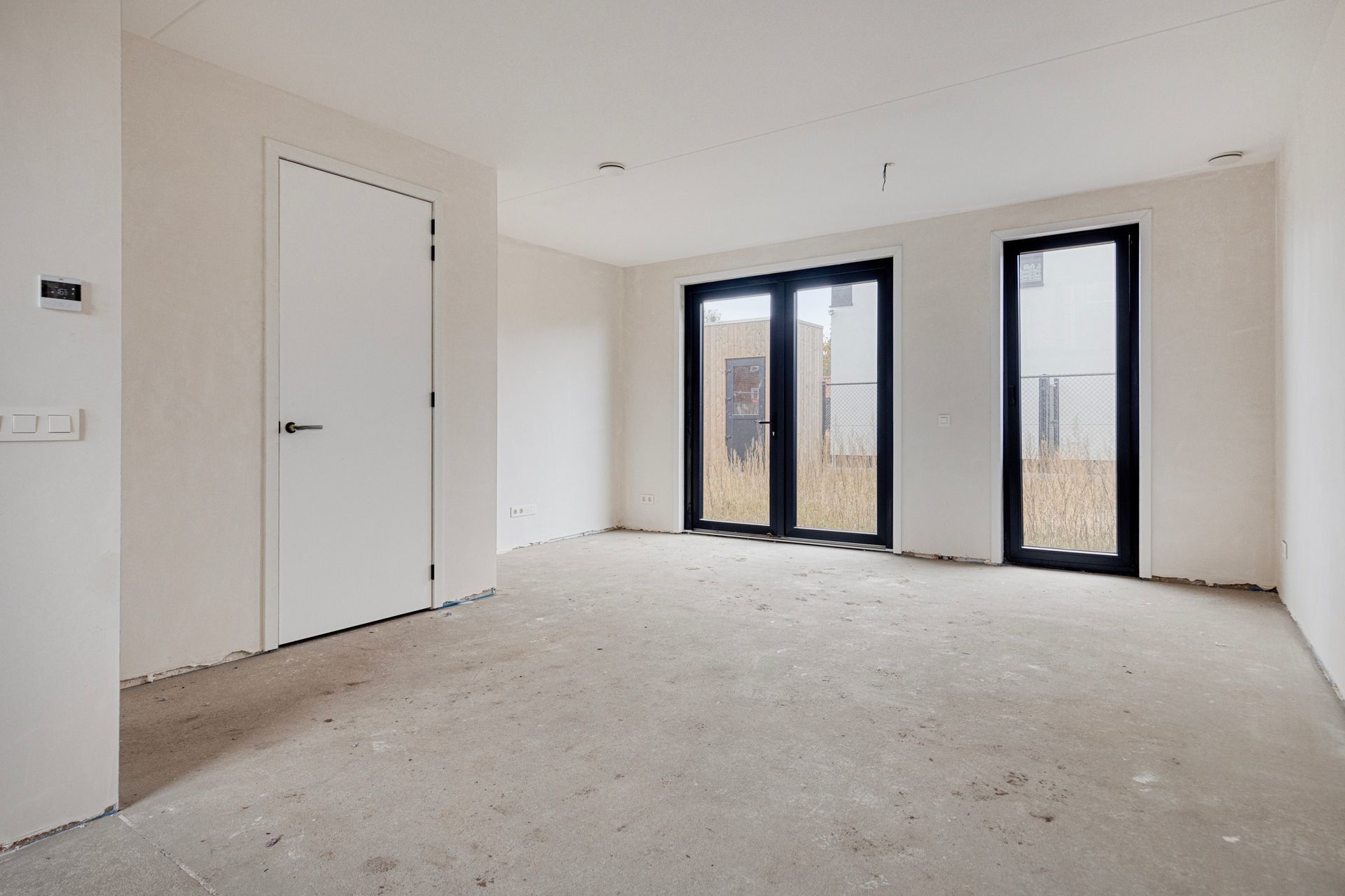 Een lege kamer met witte muren, een onafgewerkte betonnen vloer en deuren/ramen met zwarte kozijnen.