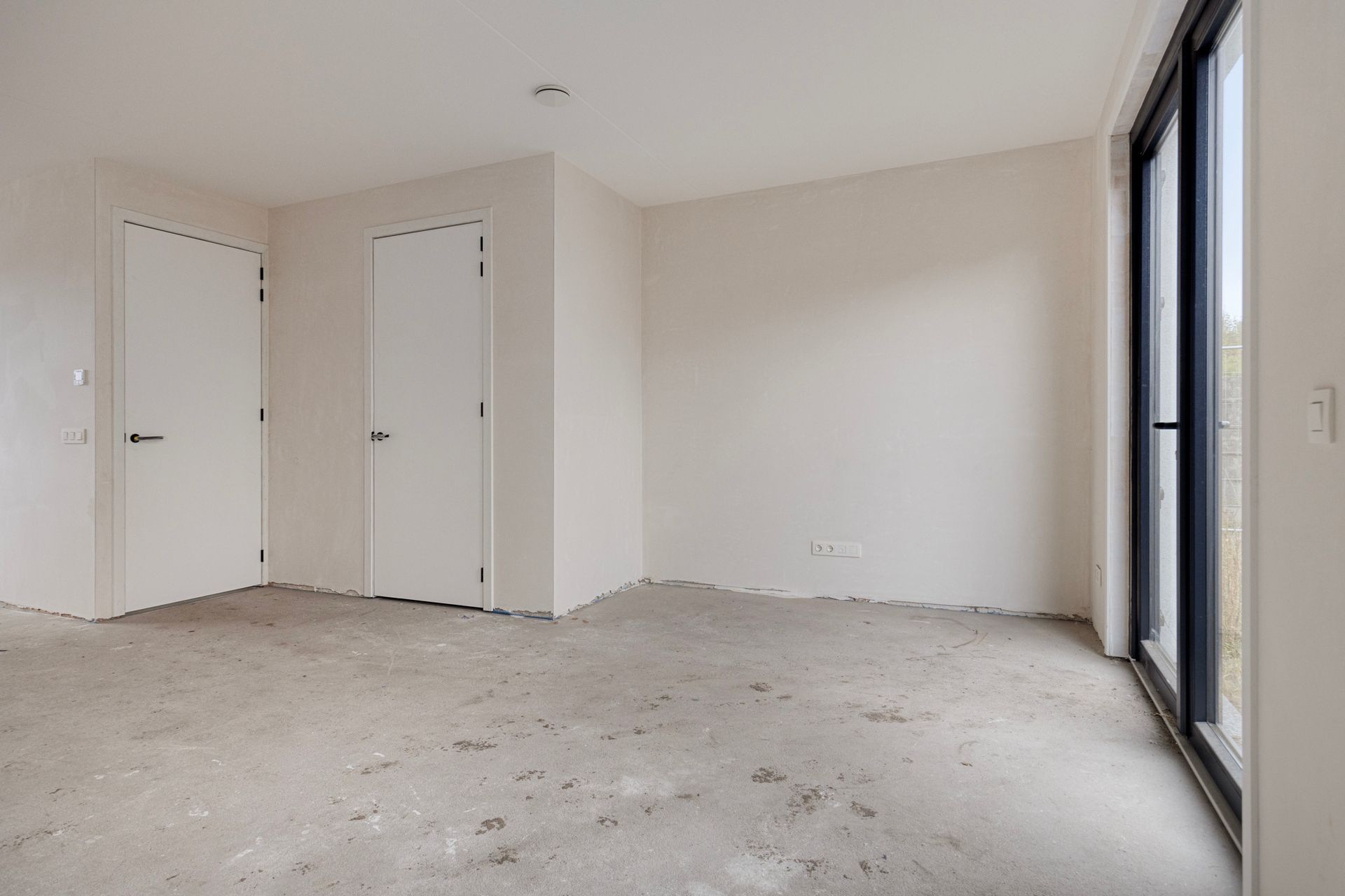 Lege binnenruimte met betonnen vloer, witte muren, twee deuren en een groot raam.