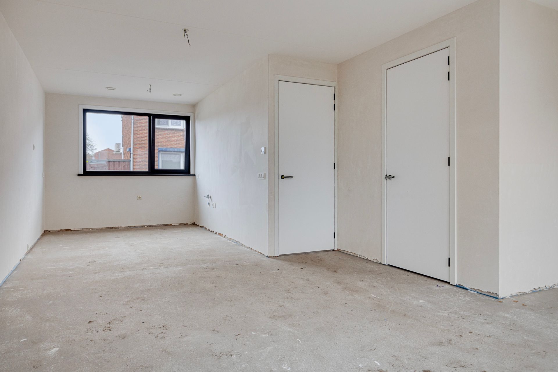 Lege kamer met onafgewerkte muren, een raam en twee gesloten witte deuren; betonnen vloer.