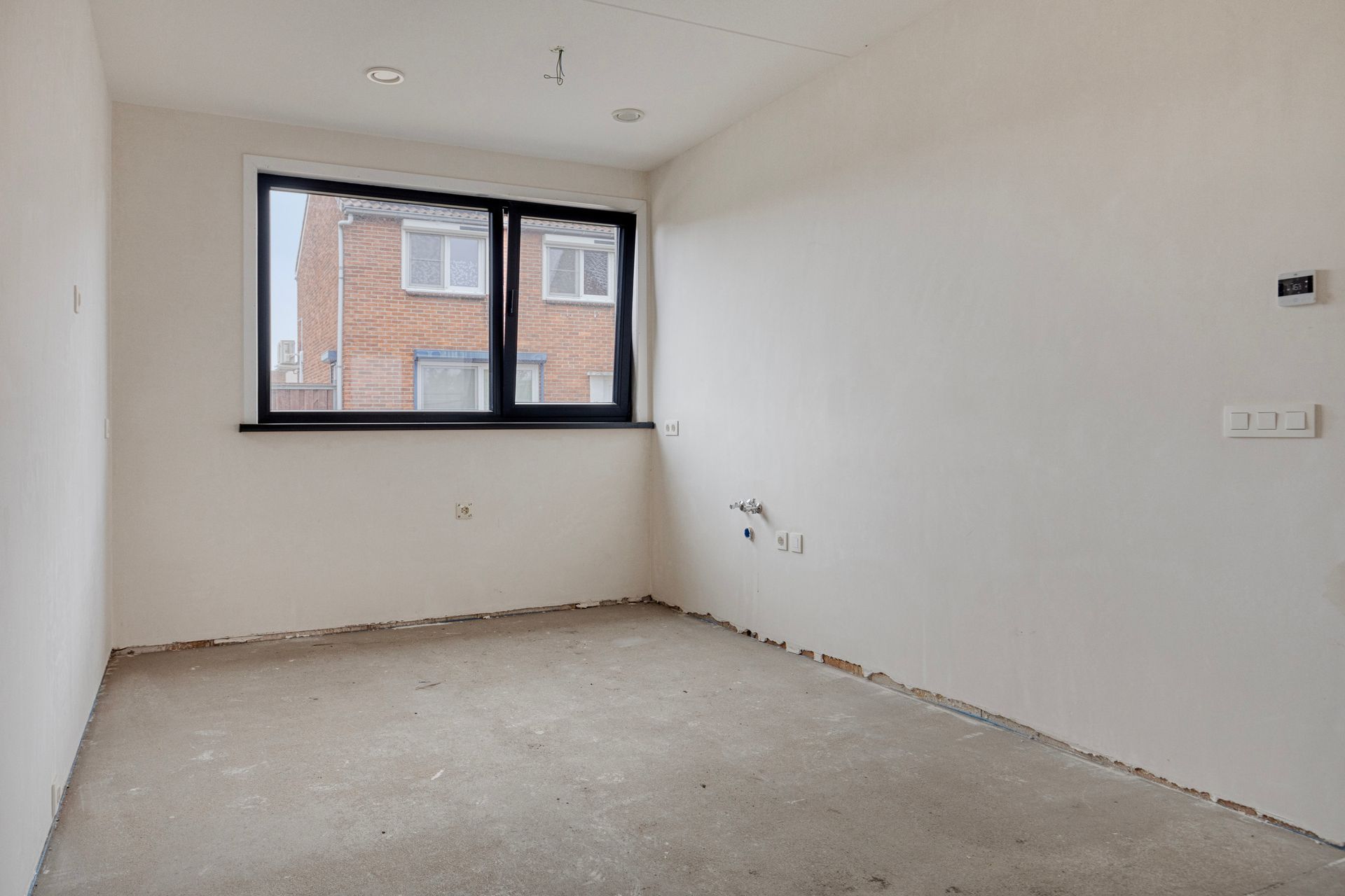 Lege kamer met raam. Witte muren, onafgewerkte vloer, stopcontacten, inbouwverlichting en uitzicht op een gebouw buiten.