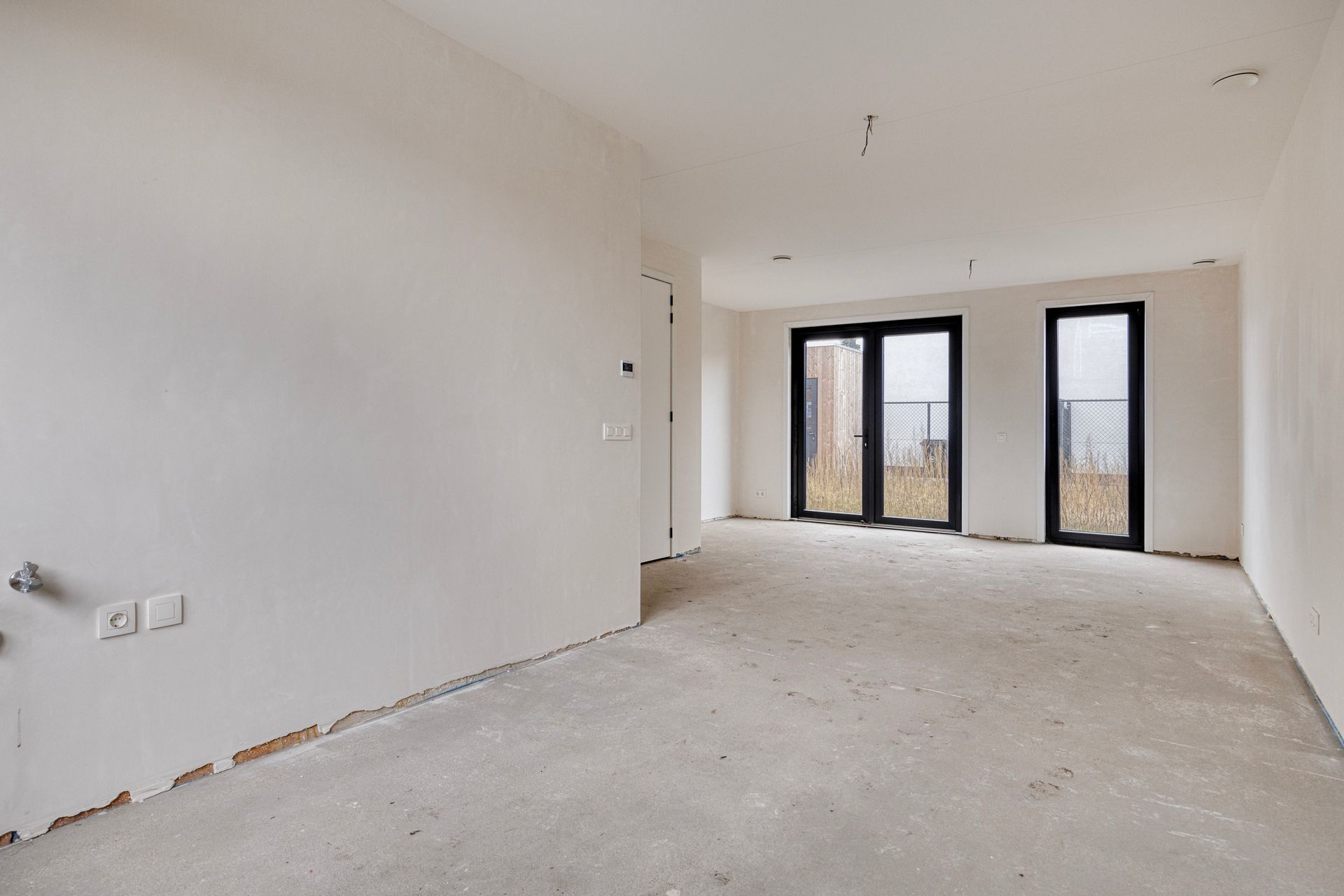 Een lege kamer met witte muren, een onafgewerkte betonnen vloer en ramen met zwarte kozijnen die uitzicht bieden op de stad.