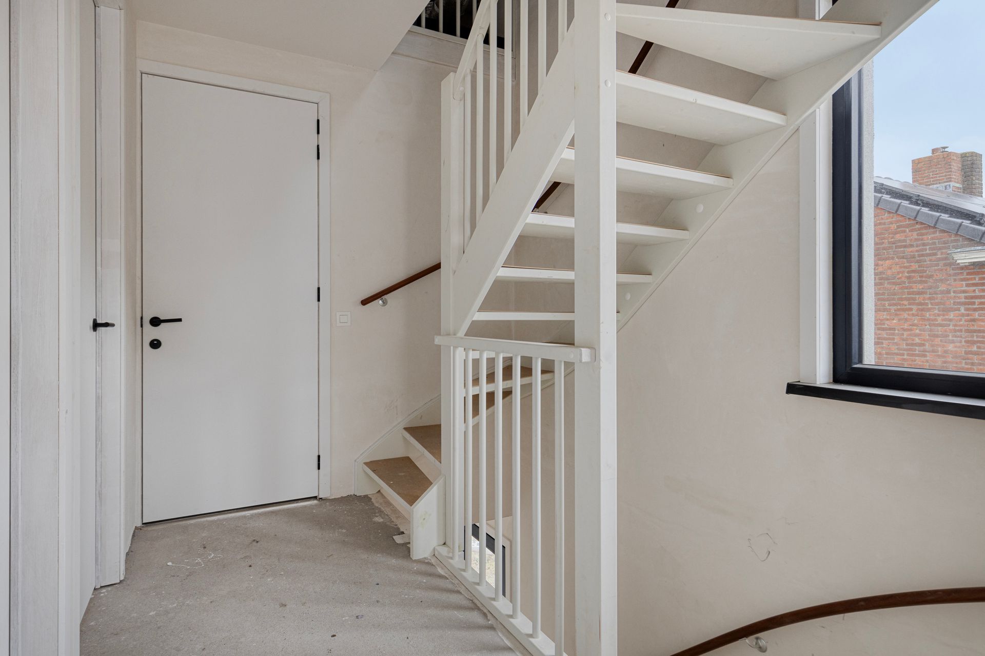 Witte trap met houten treden, die naar boven leidt. Witte deur en raam zichtbaar.