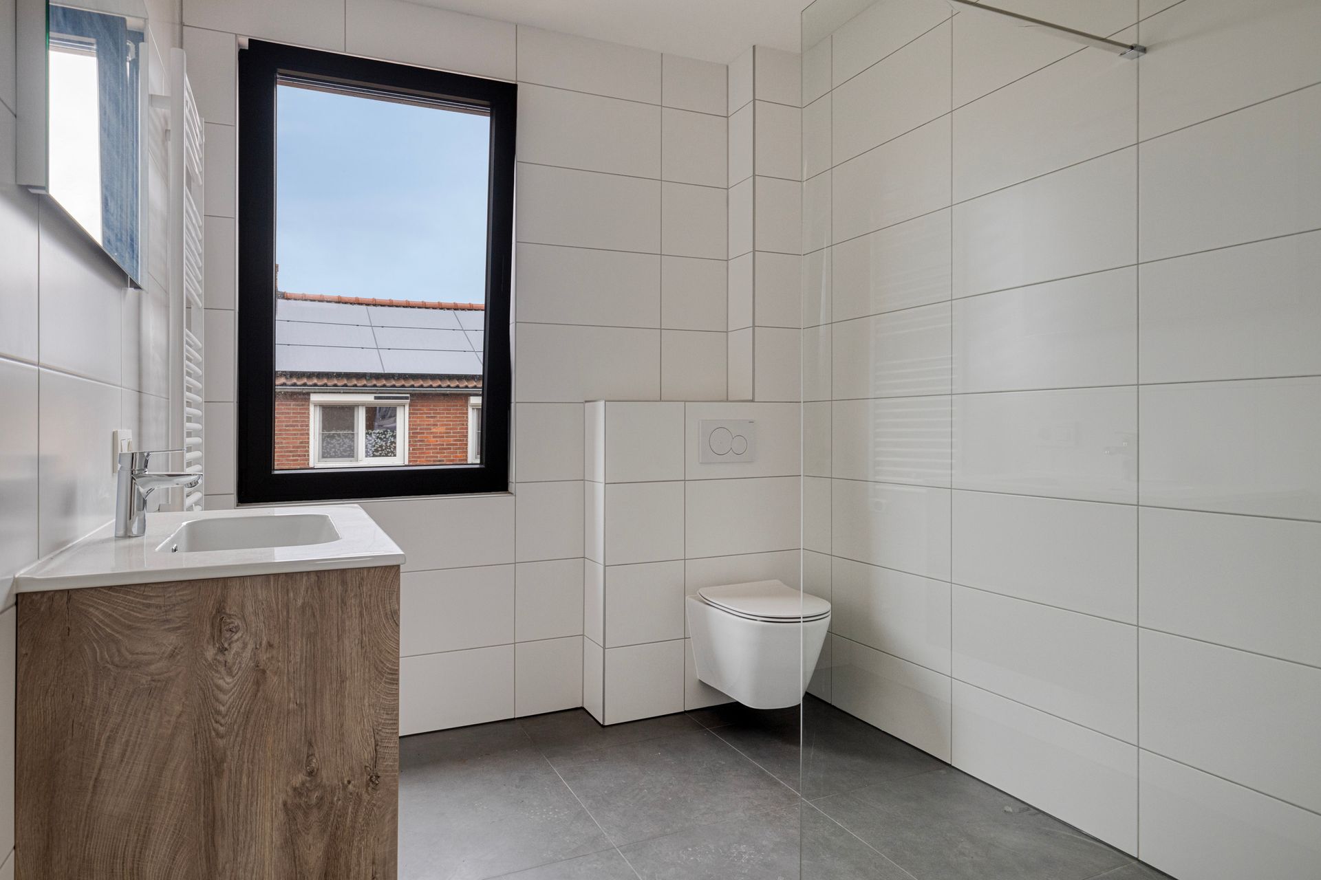 Badkamer met witte tegels, een raam met uitzicht op de daken, een zwevend toilet en een houten wastafelmeubel.