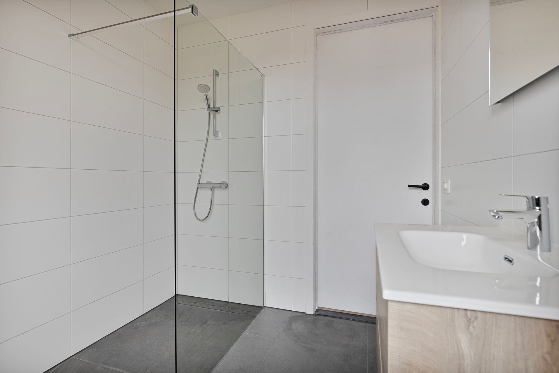 Witte badkamer met een glazen douchecabine, een wastafel met een houten kastje en een gesloten witte deur.