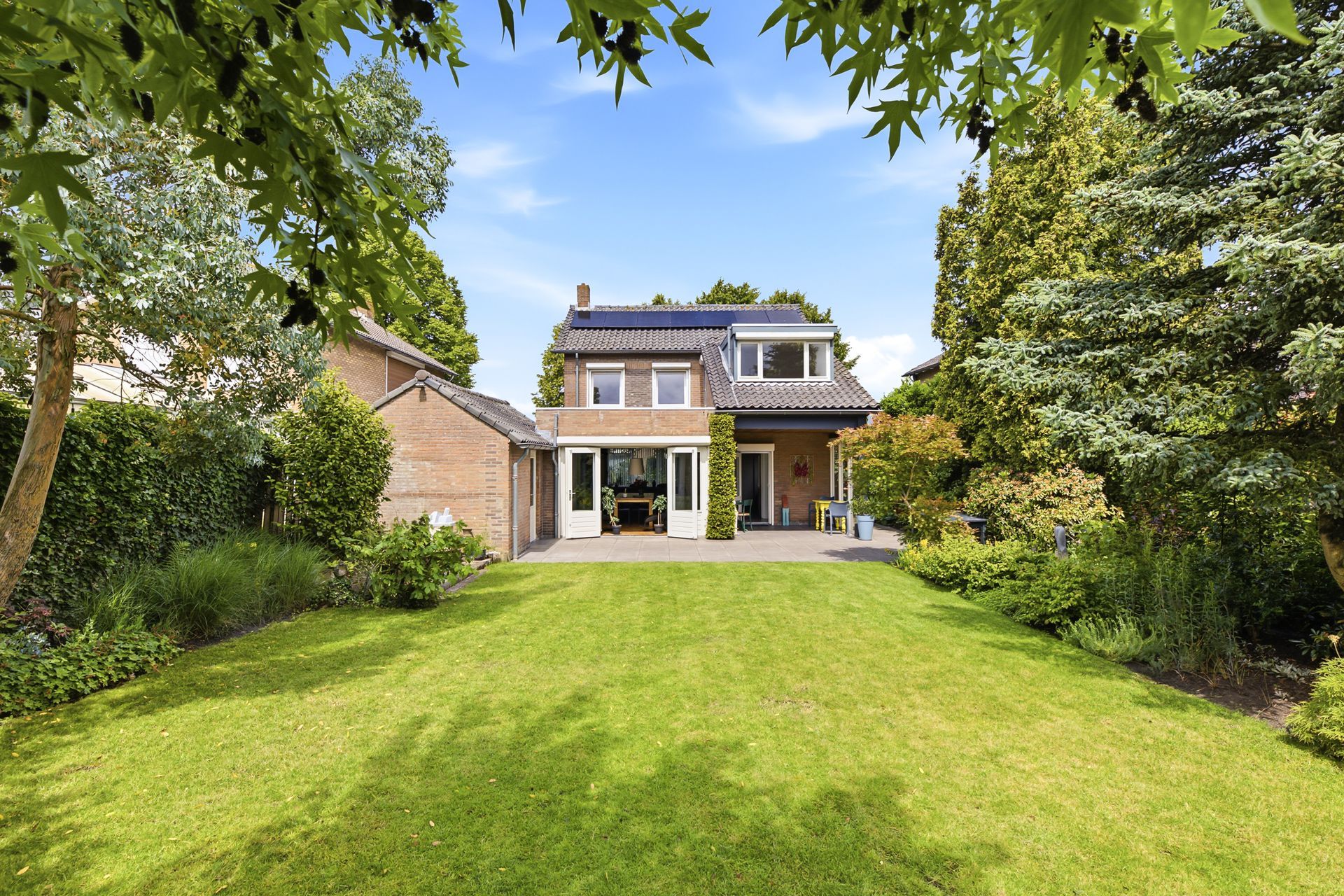 Terras in de achtertuin met bestrating, groen gazon en uitzicht op de huizen van de buren onder een blauwe hemel.