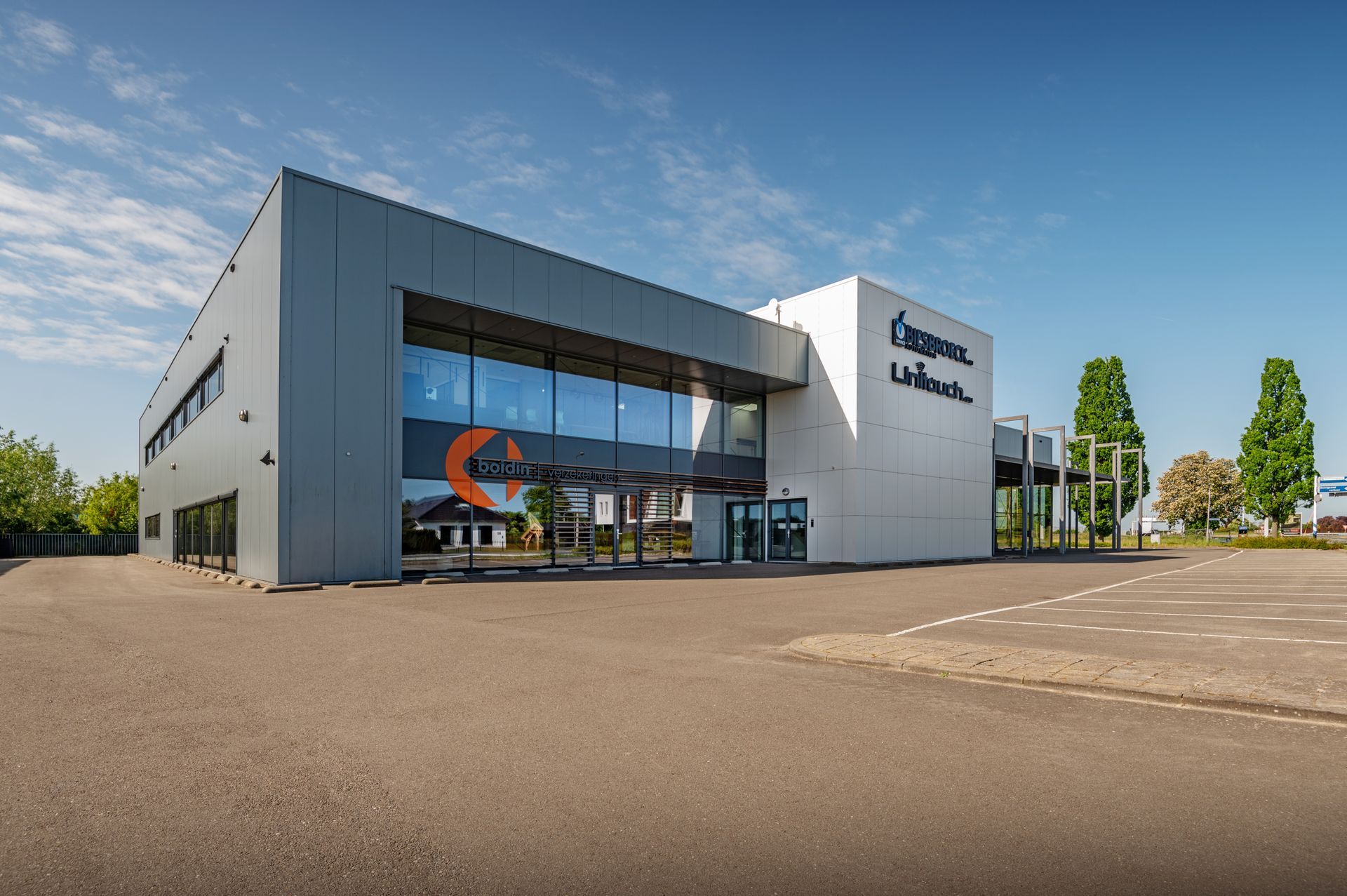 Modern grijs gebouw met grote ramen en een oranje logo. Verharde parkeerplaats ervoor.