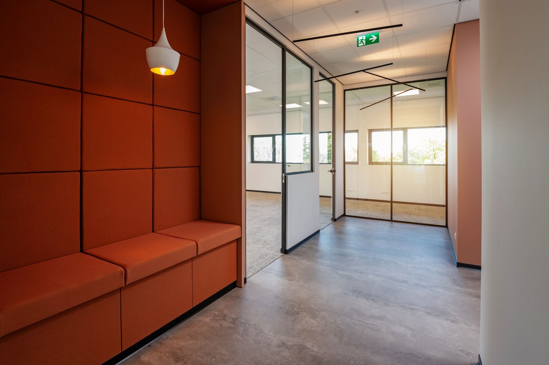 Gang met oranje zitmeubels en wandpanelen, die leidt naar kantoren met glazen wanden.