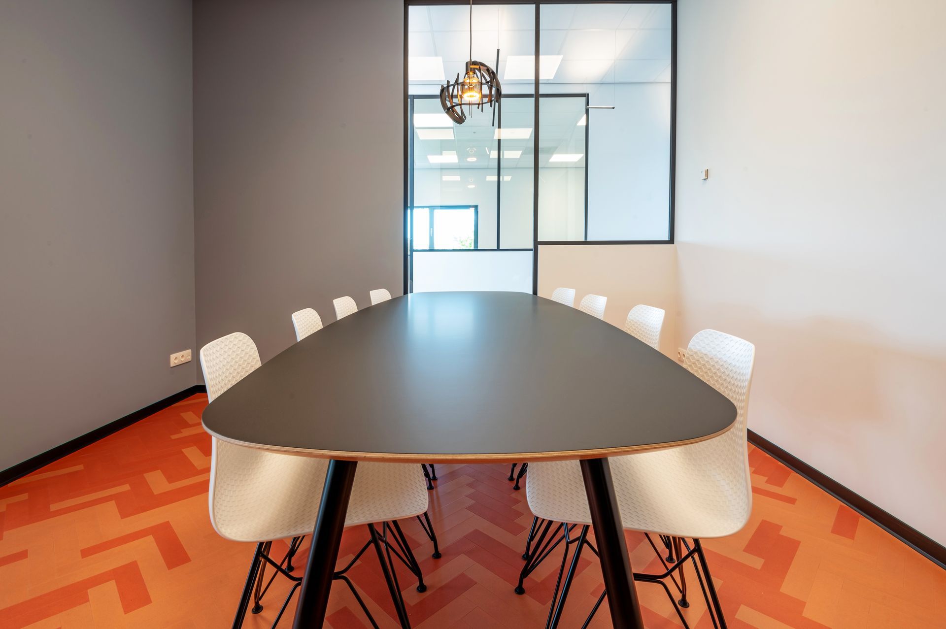 Moderne vergaderruimte met driehoekige tafel, witte stoelen, oranje vloer en glazen wand.