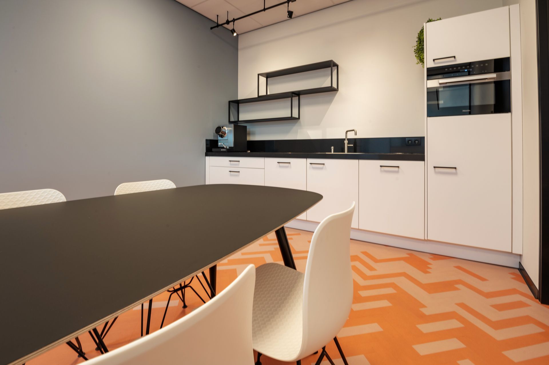 Kitchenette met een lange tafel en stoelen; witte kasten, zwart aanrechtblad, oranje vloer.