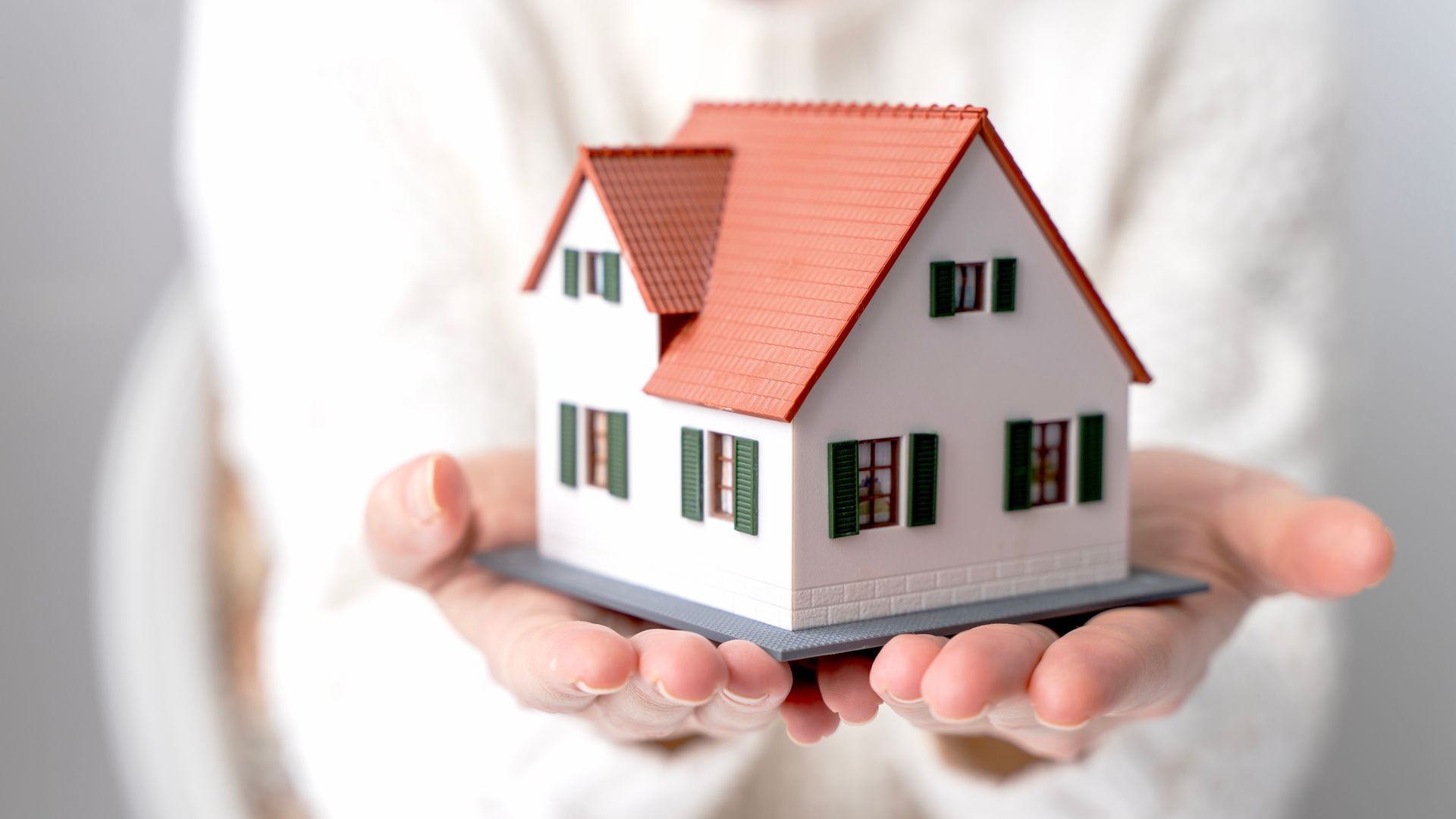 Een persoon houdt een klein modelhuisje vast met een oranje dak, witte muren en groene luiken.