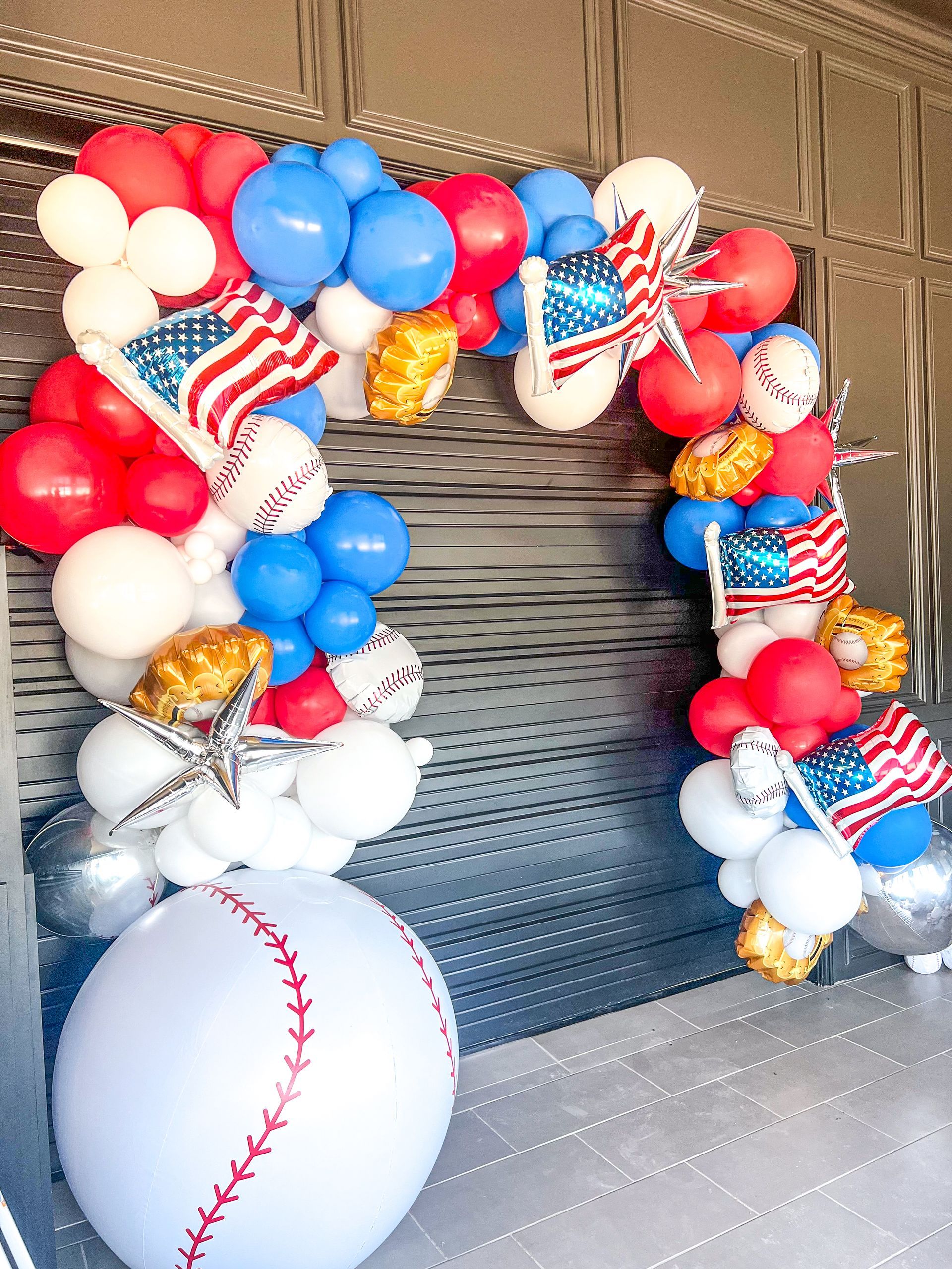 A red , white , and blue balloon arch with a baseball in the middle.