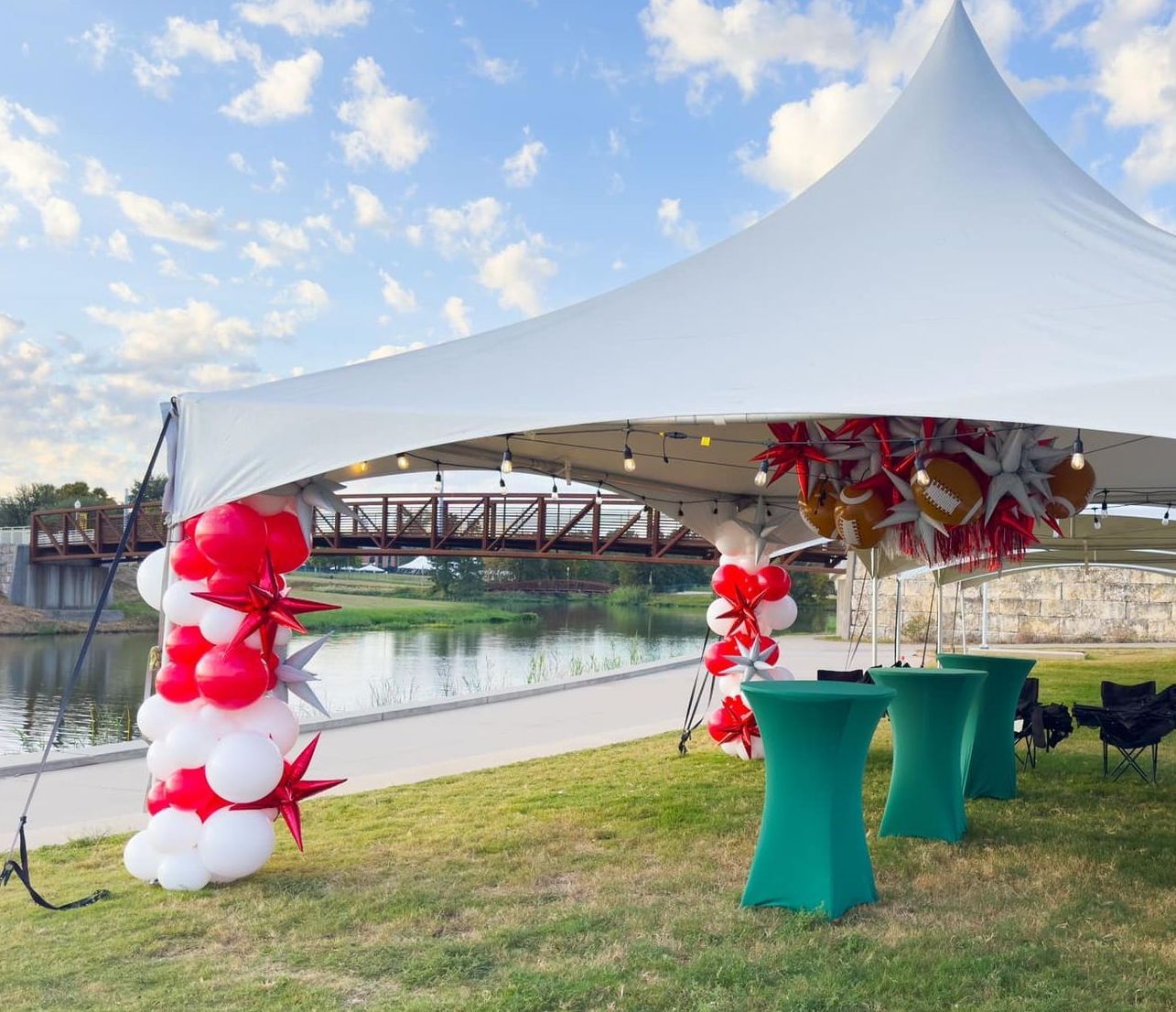 A white tent is decorated with red and white balloons and green tables.