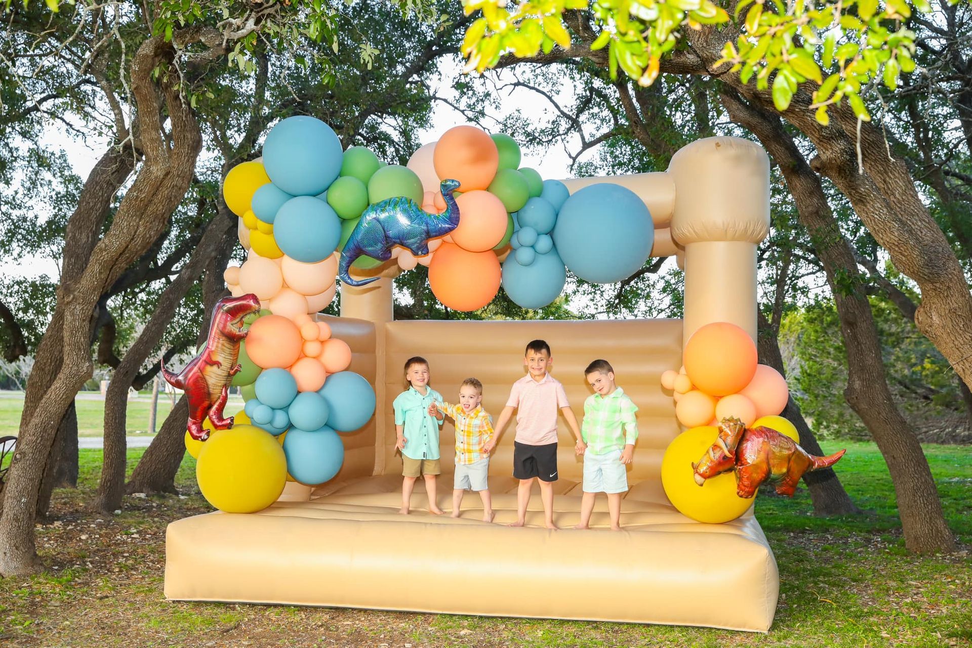 A group of children are standing in front of a bouncy house decorated with balloons and dinosaurs.