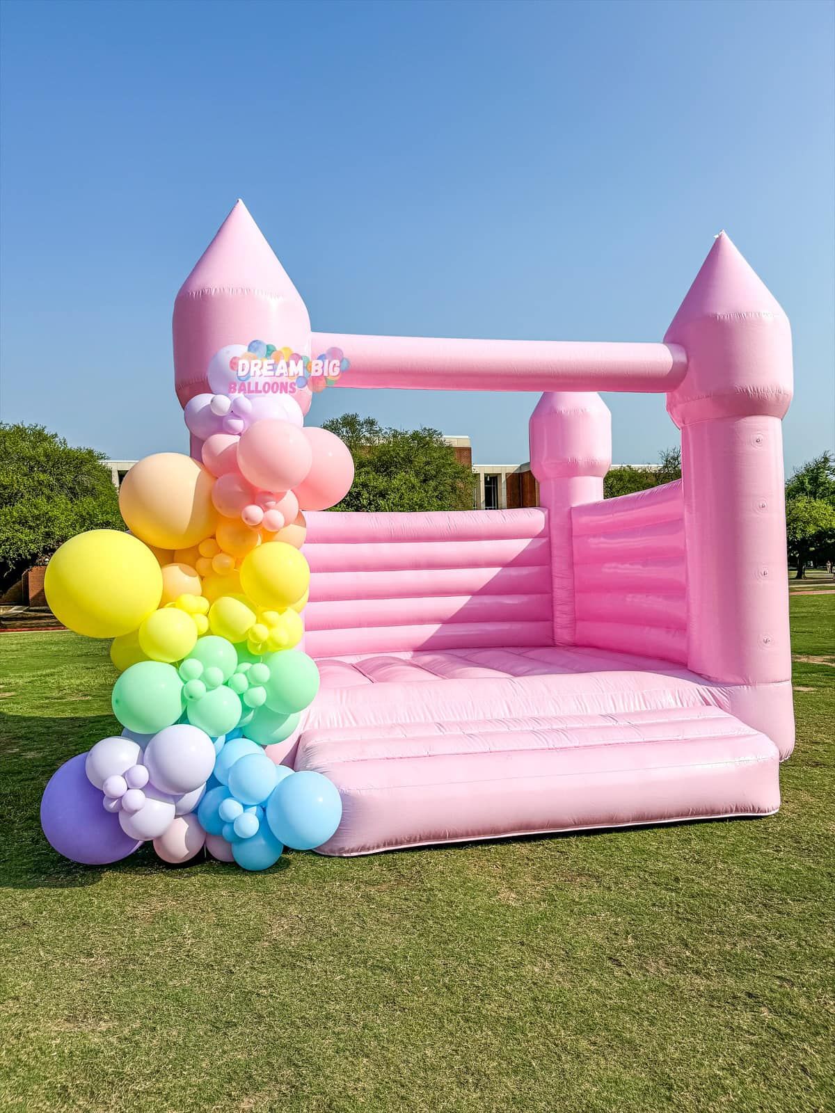 A pink bouncy castle surrounded by balloons on a lush green field.