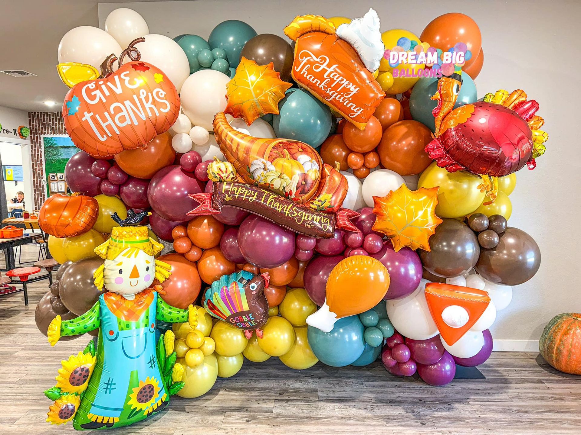 A scarecrow is standing in front of a wall filled with balloons for thanksgiving.