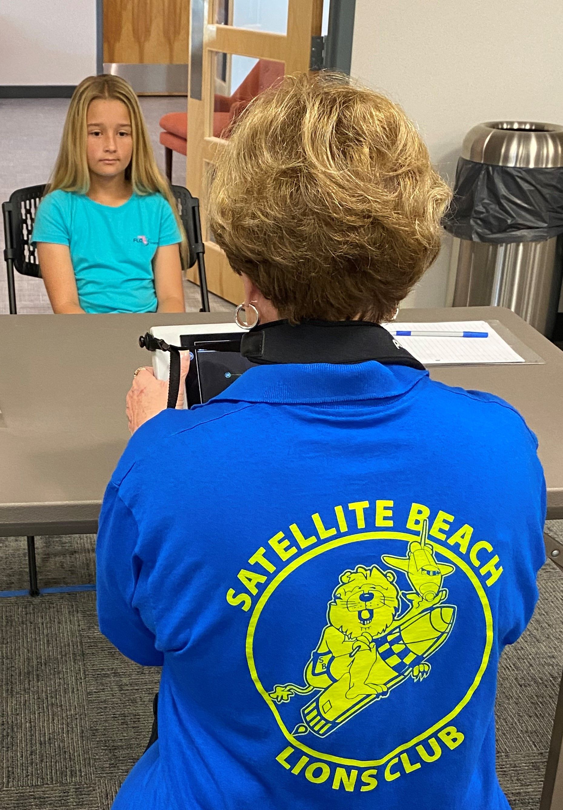 A woman wearing a blue satellite beach lions club jacket is taking a picture of a girl sitting at a table.