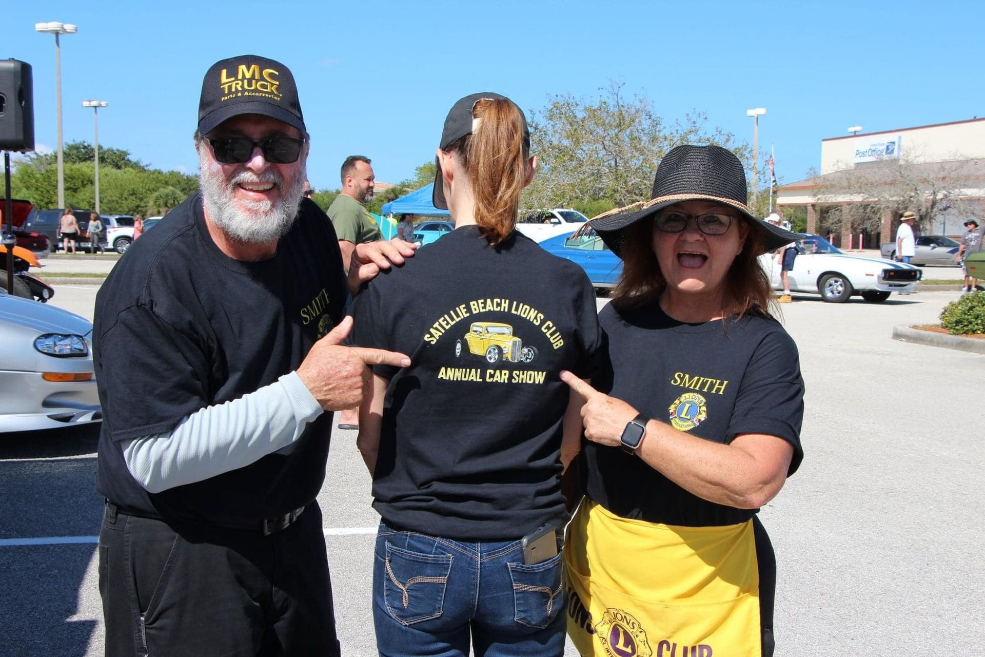 A man and two women are standing in a parking lot pointing at their shirts