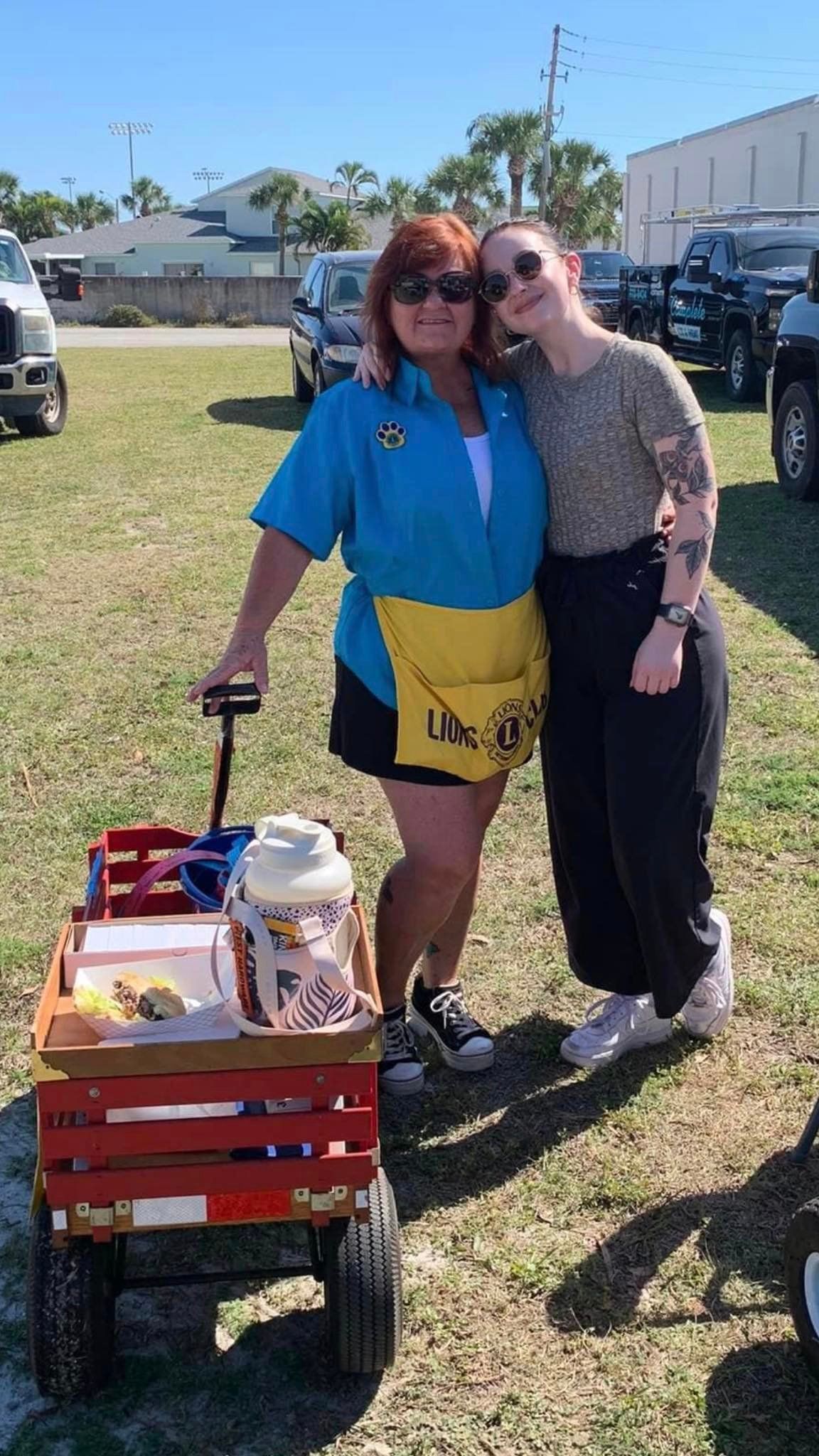 Two women are standing next to a red wagon in a field.