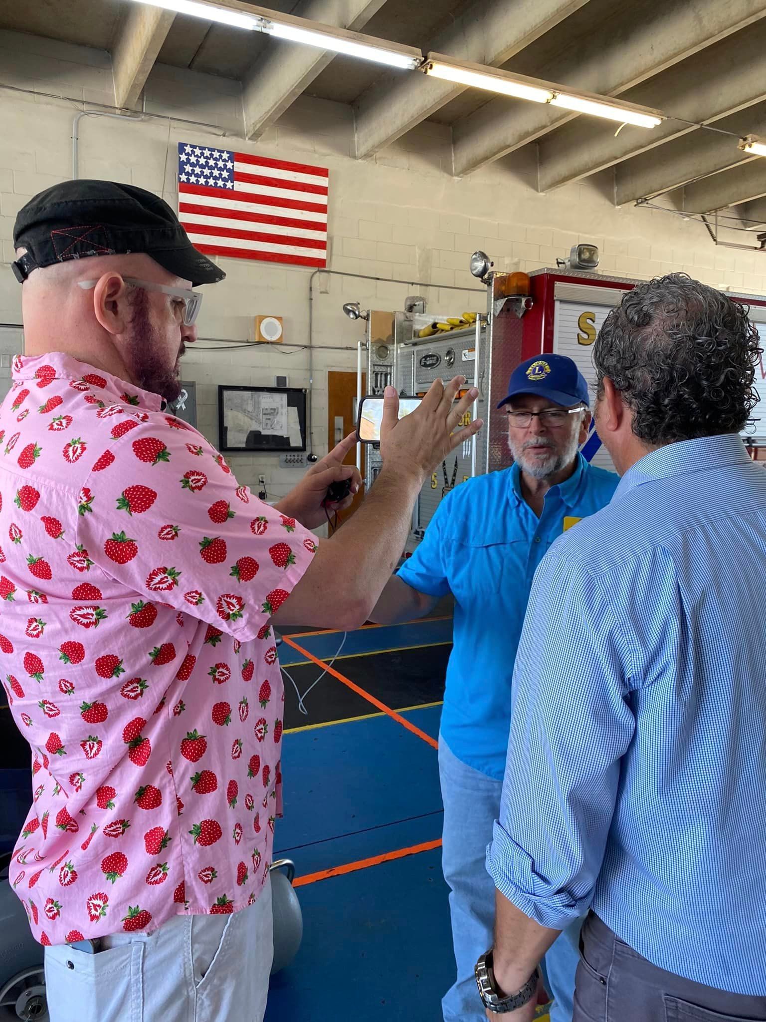A man in a pink shirt with strawberries on it is talking to two other men in a room.