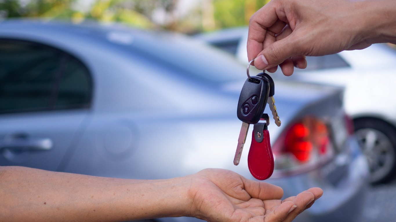 No primeiro plano da imagem aparece a mão de um homem entregando as chaves de um carro para uma mão feminina. Em segundo plano há dois carros estacionados.