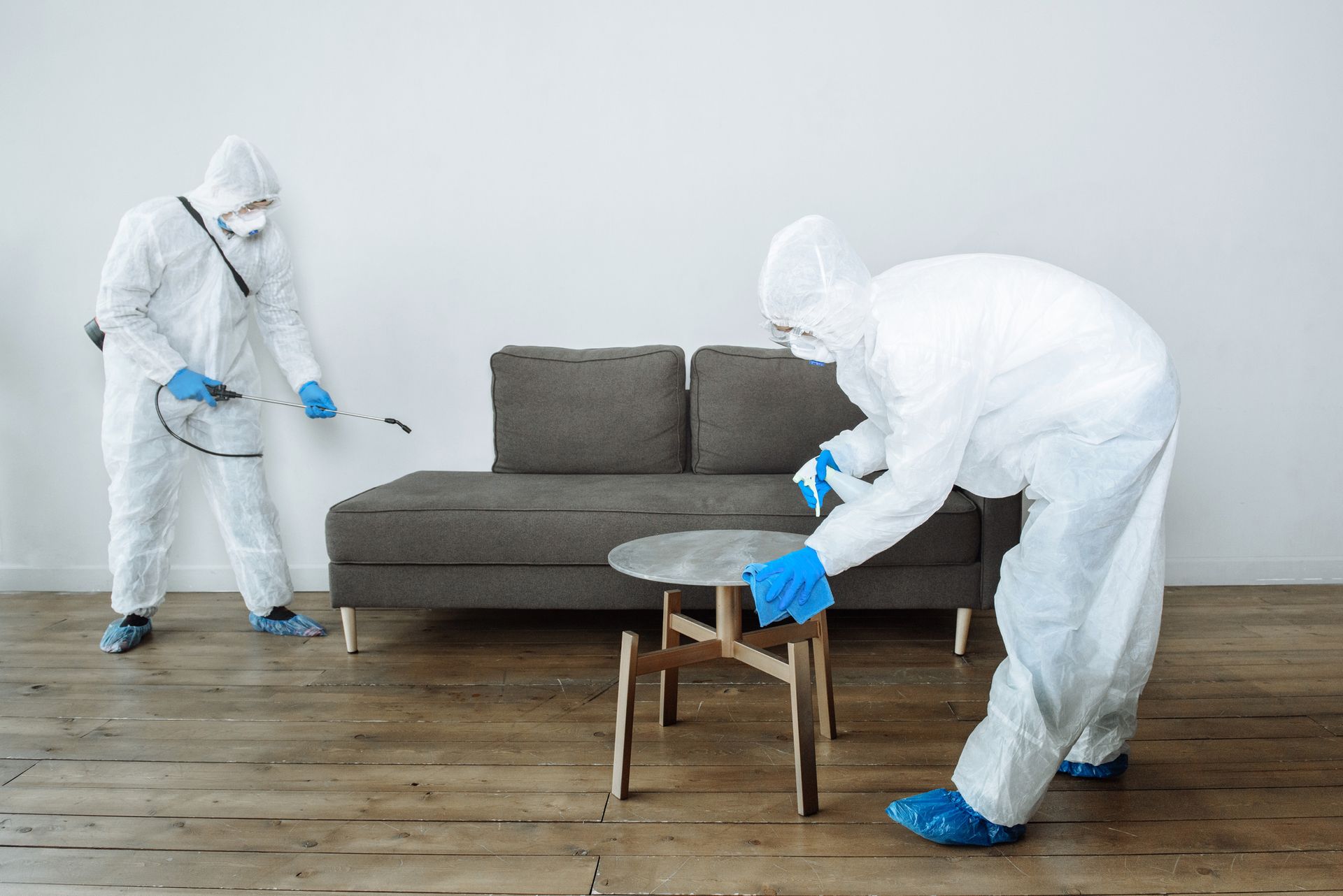 Two individuals in full protective suits and gloves disinfecting a living room.