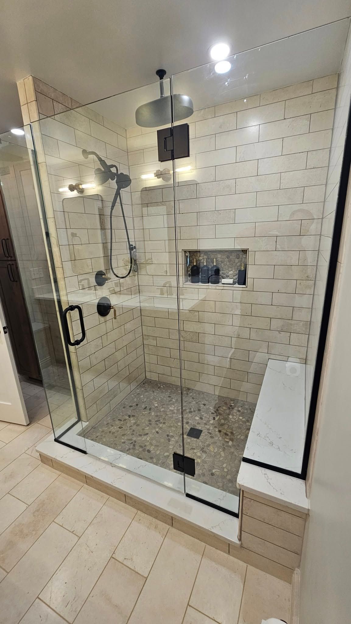 Glass-enclosed shower with a bench and stone floor. Beige tile walls. Black shower fixtures.