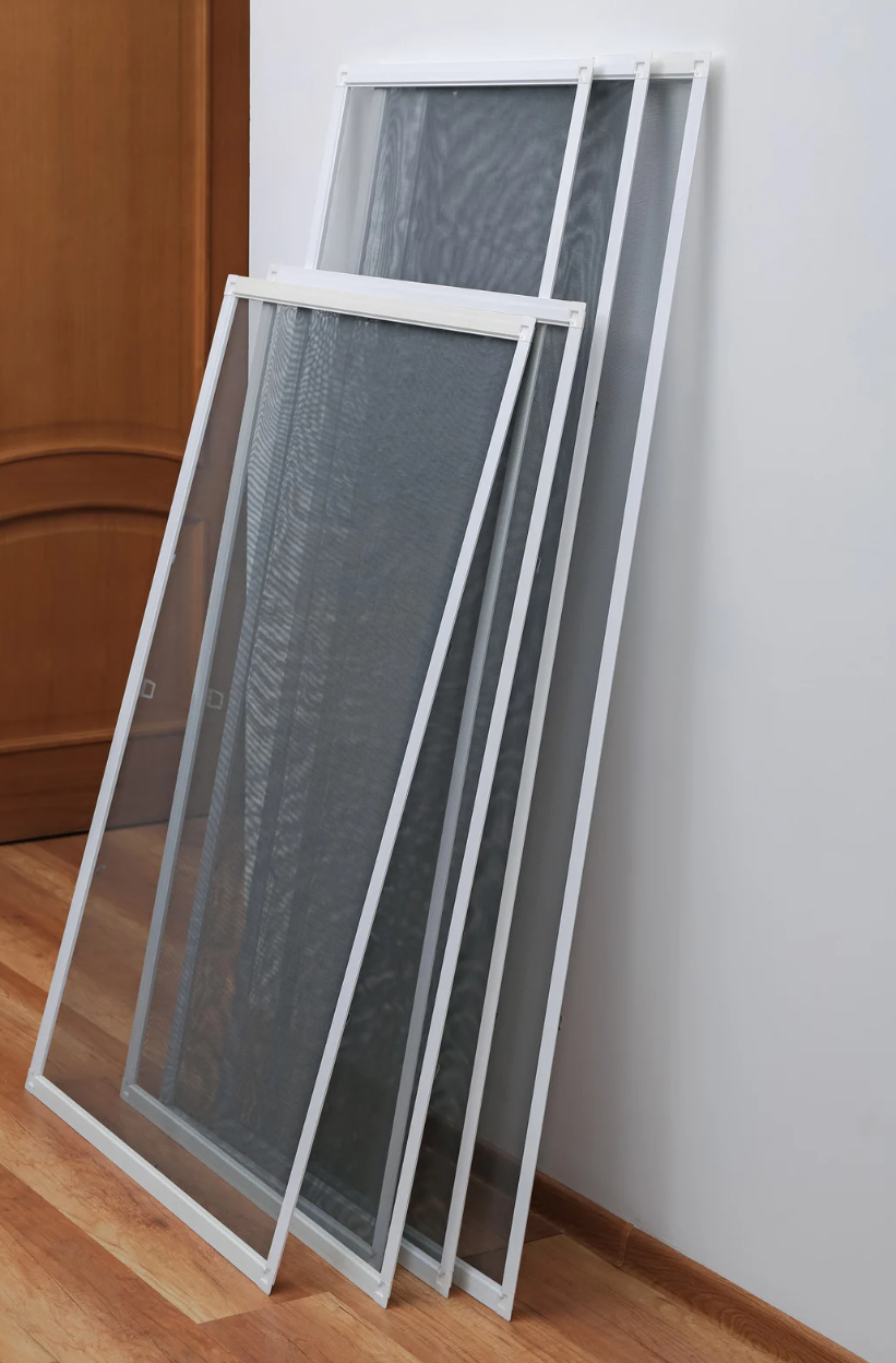 White-framed window screens leaning against a white wall and a brown door on a wood floor.