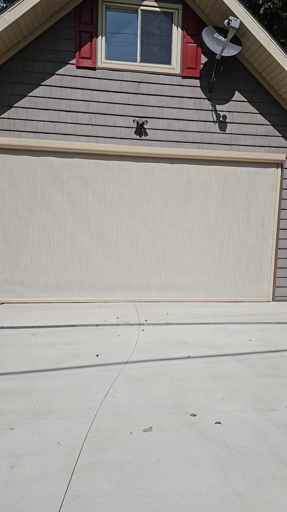 Gray garage with beige door, window, red shutters, and concrete driveway. A drone is hovering.