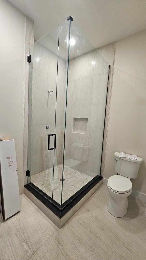 Glass shower enclosure with black hardware in a bathroom with neutral-toned tiling and a toilet.