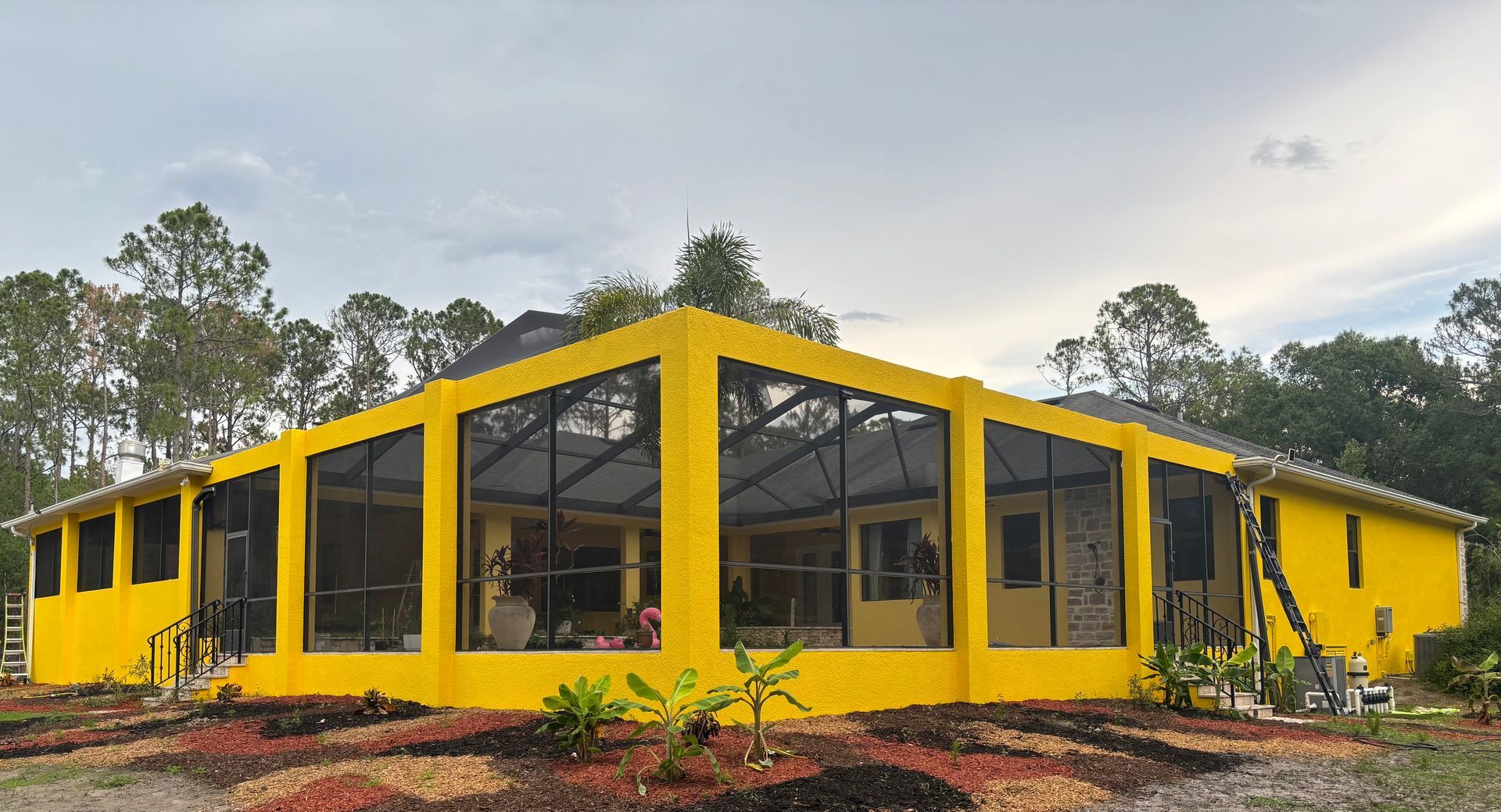 Yellow house with black screened windows, surrounded by landscaping under a cloudy sky.