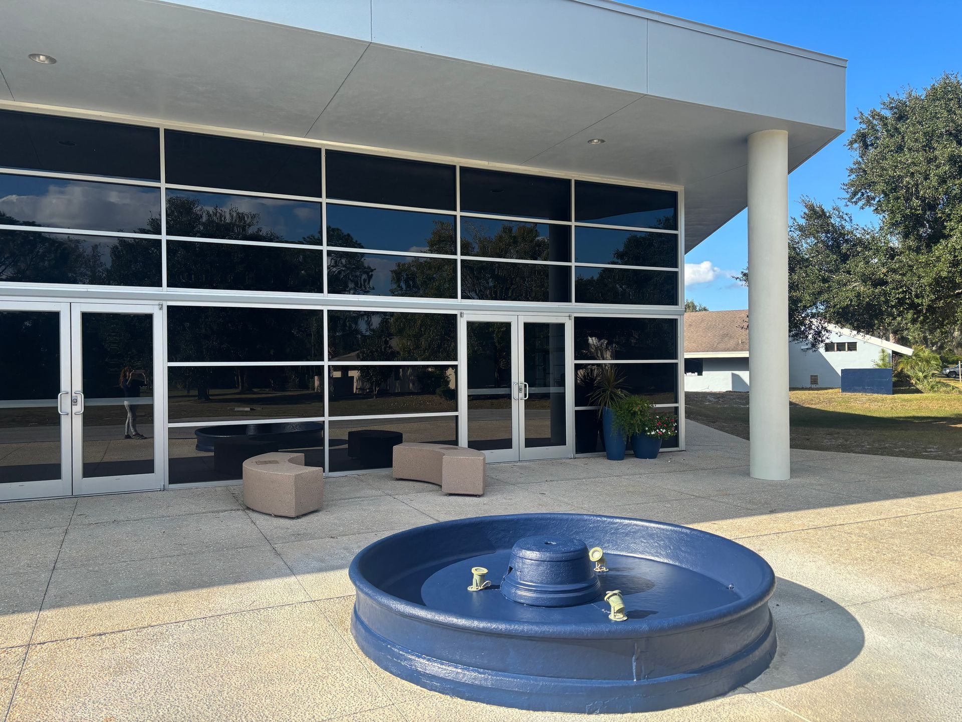 Modern building with glass windows, a blue fountain, and seating area. Sunny day.