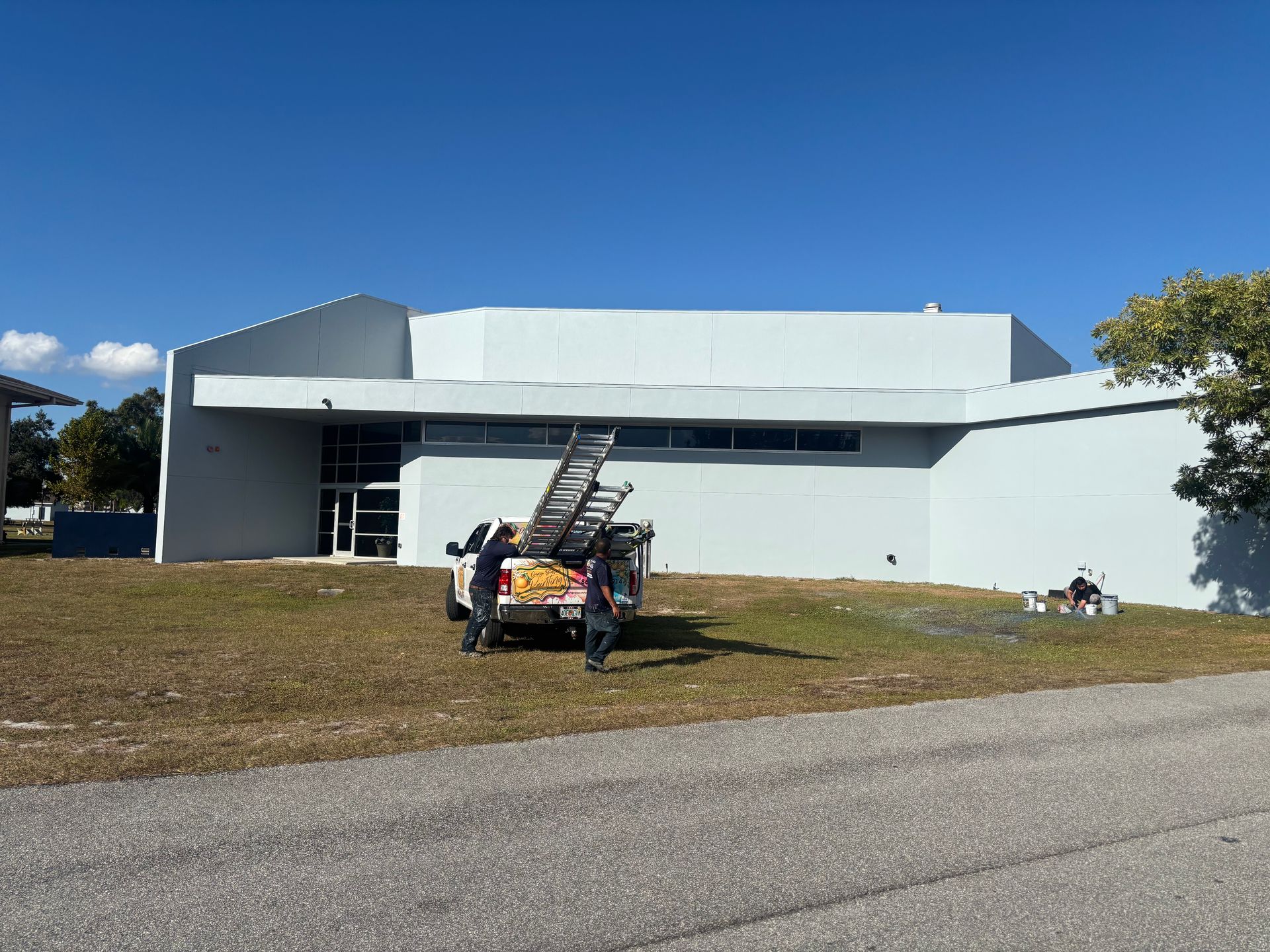 A light blue building with a maintenance truck parked in front. Two people are near the truck, and a ladder is leaning on it.