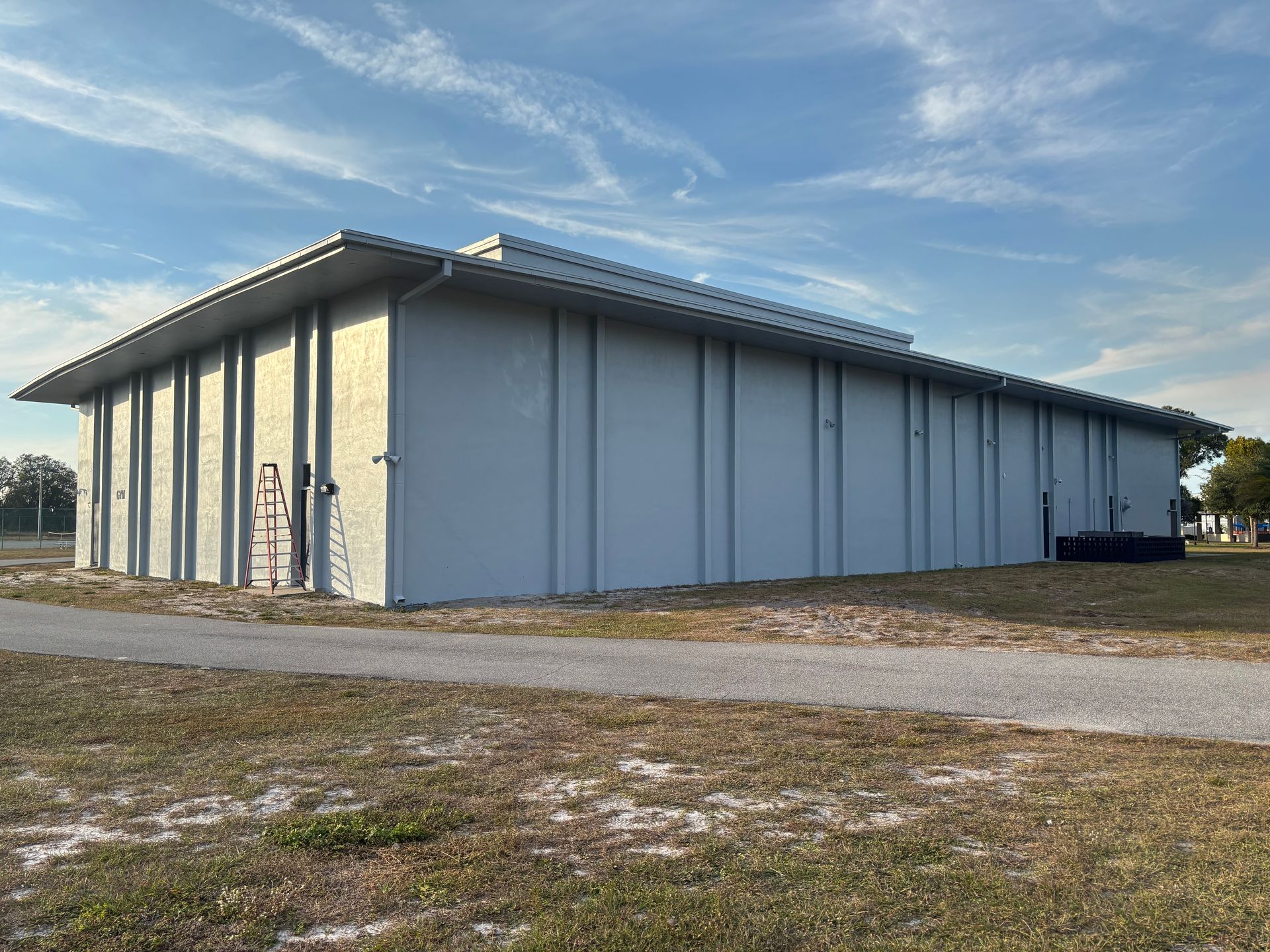 A light gray industrial building with a flat roof on a grassy field, under a partly cloudy sky.