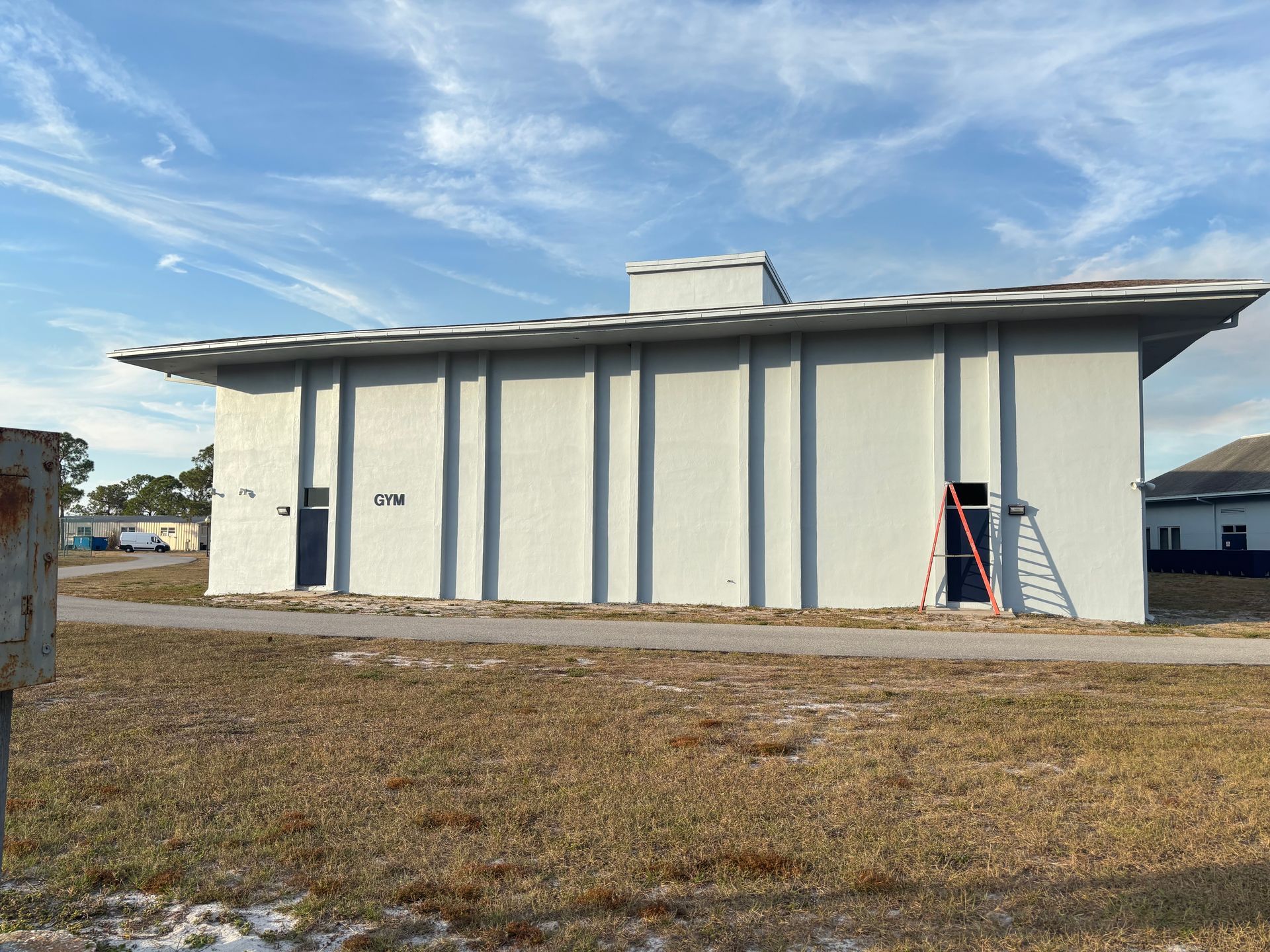Gray building with dark blue doors, ladder, and a flat roof against a partly cloudy sky.