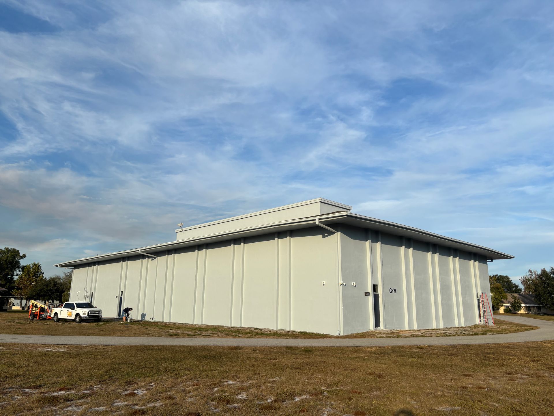 Gray building with a flat roof under a blue sky; a white truck is parked nearby.