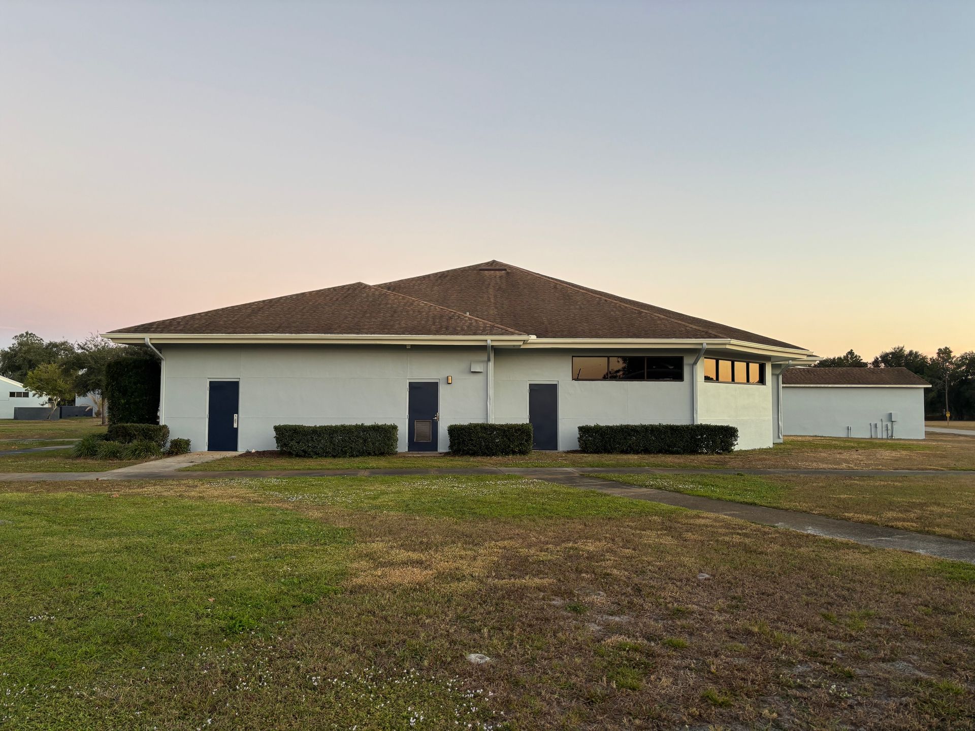 White building with brown roof, blue doors, and short green hedges. Lawn in front under a sunset sky.