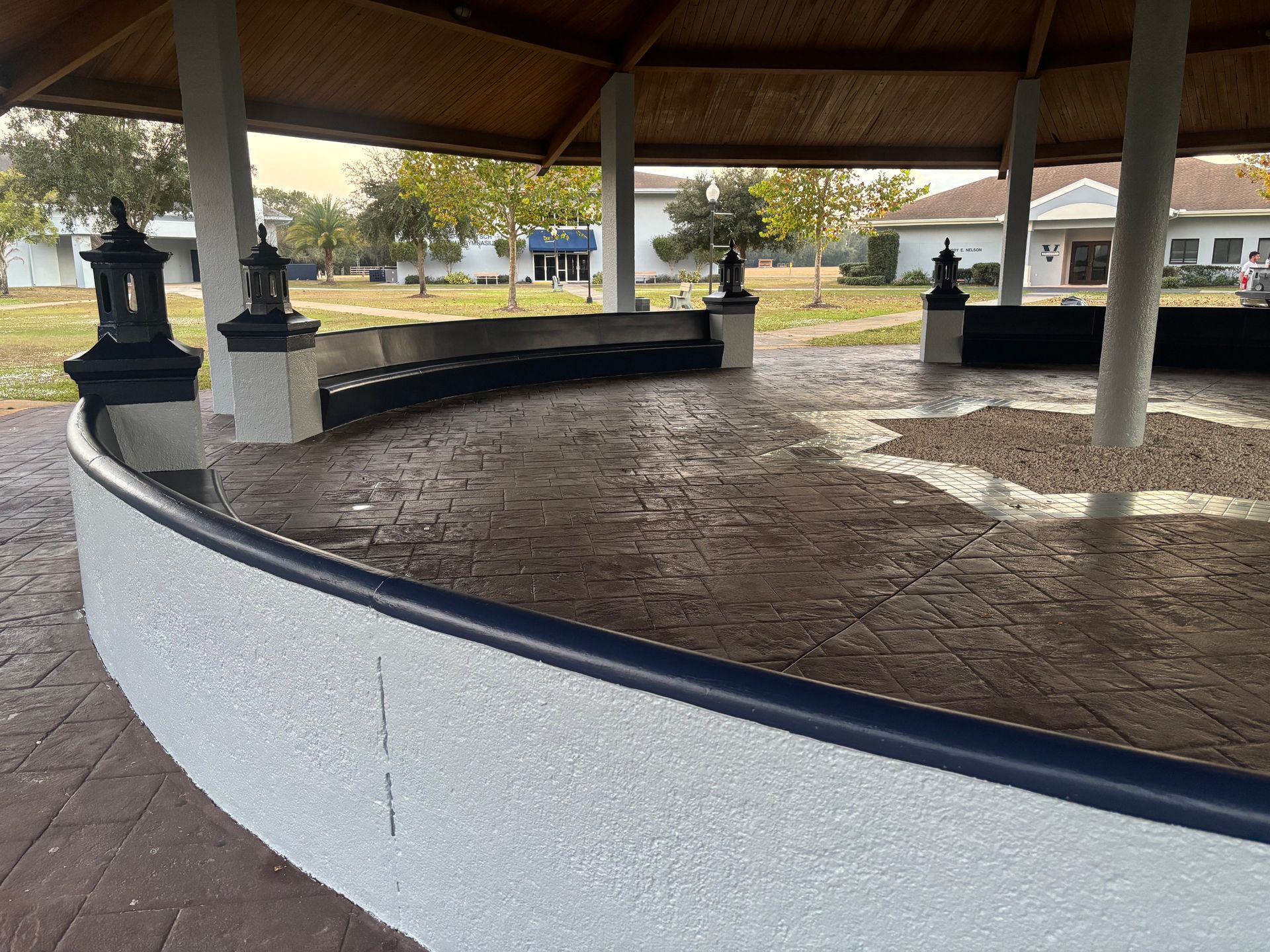 Gazebo with curved bench seating, decorative lamps, and textured floor, outdoors.