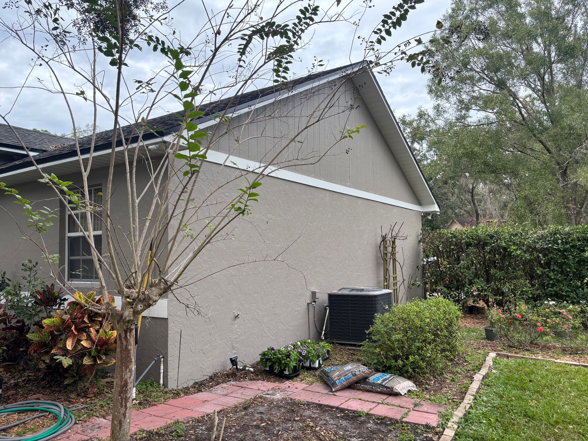 Tan house with white trim, AC unit, and landscaping under a cloudy sky.