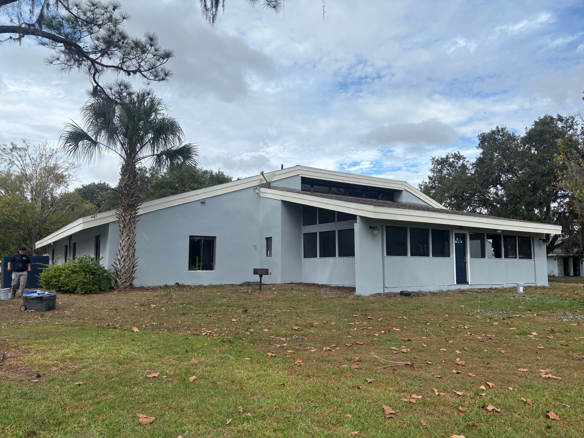 A gray, single-story building with a screened porch and a partially damaged roof, set on a grassy lot.
