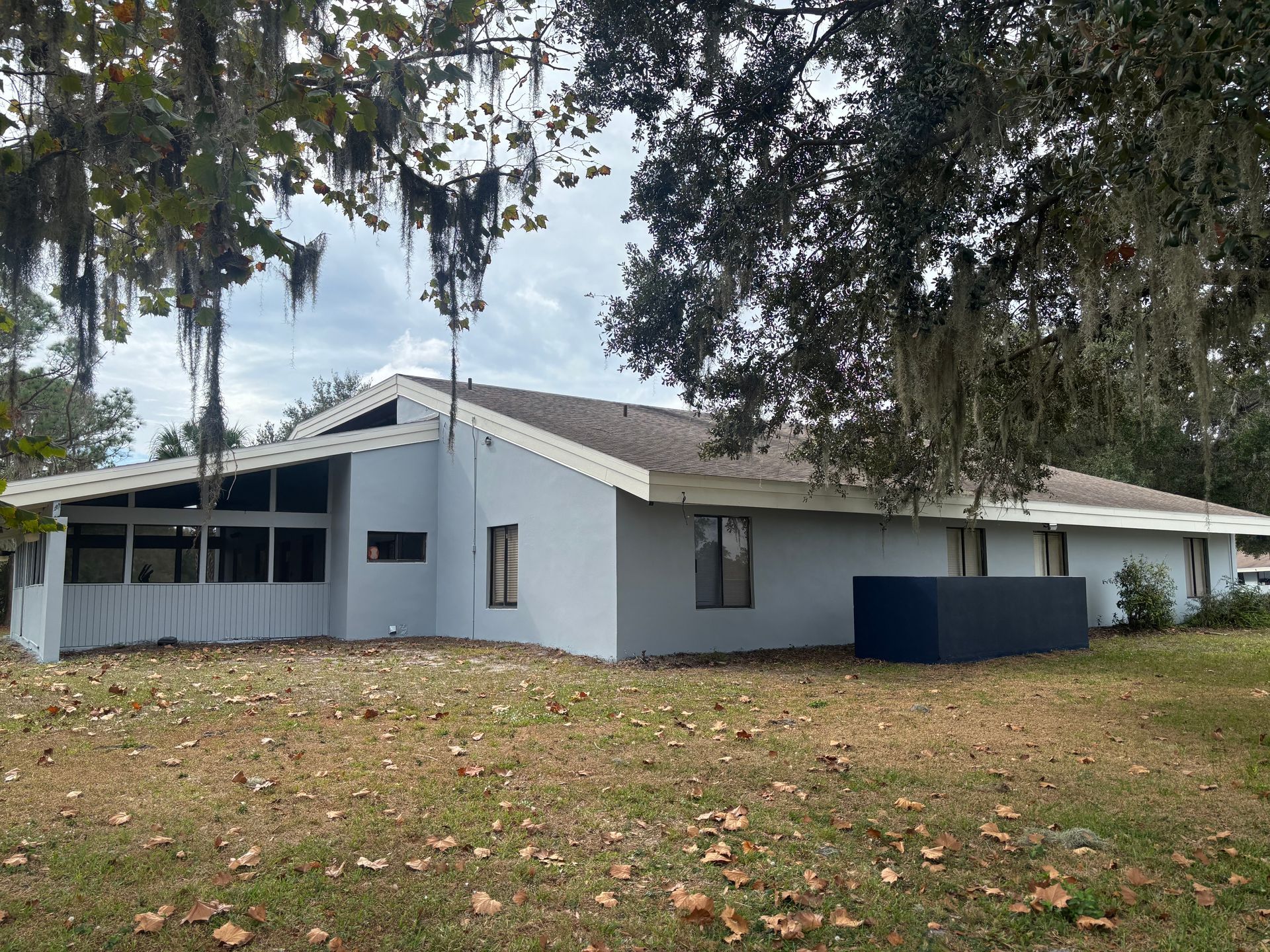 Gray house with porch, under trees with Spanish moss, on grassy lawn. Cloudy sky.