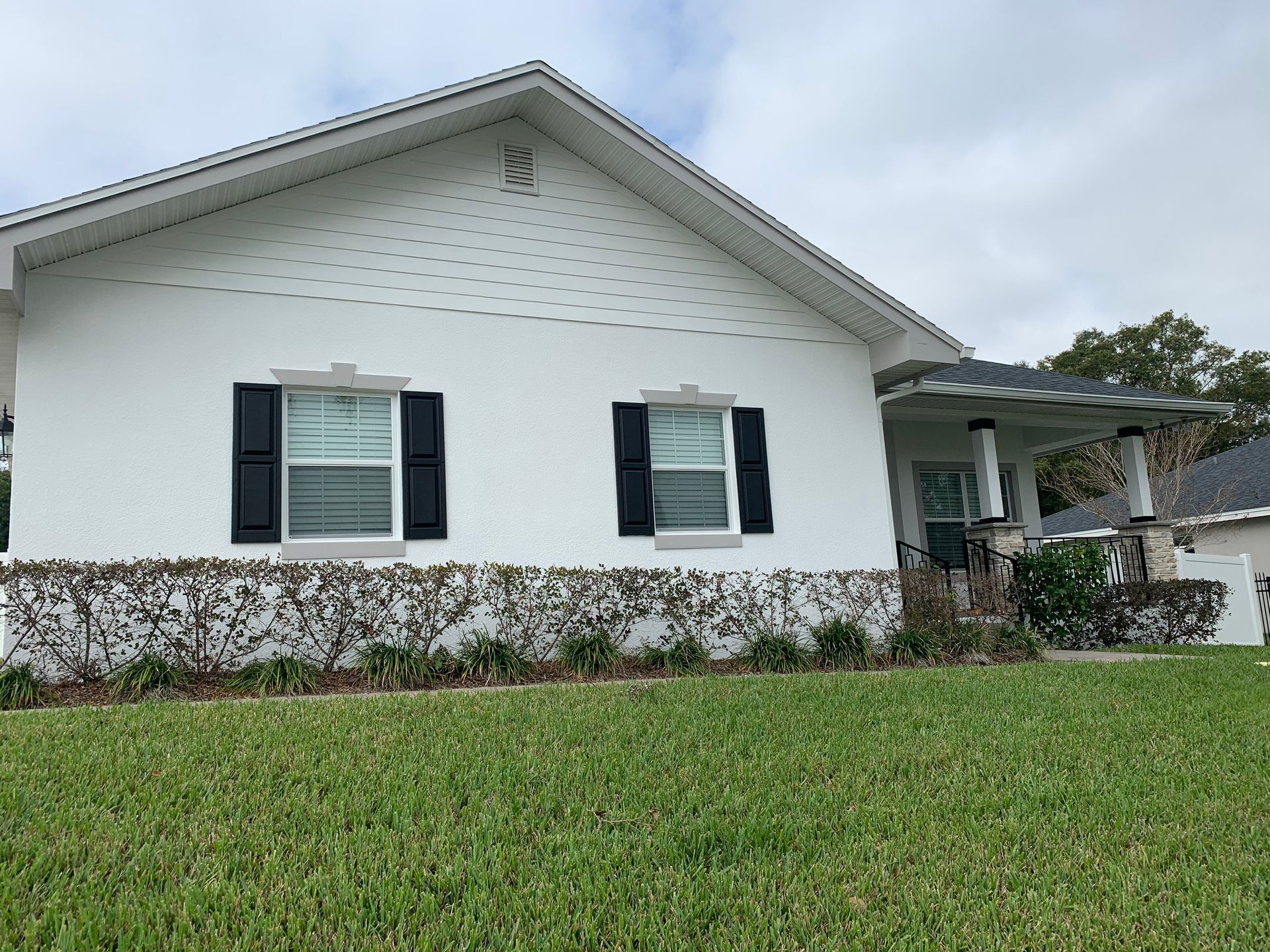 White stucco house with black shutters, green lawn, and cloudy sky.