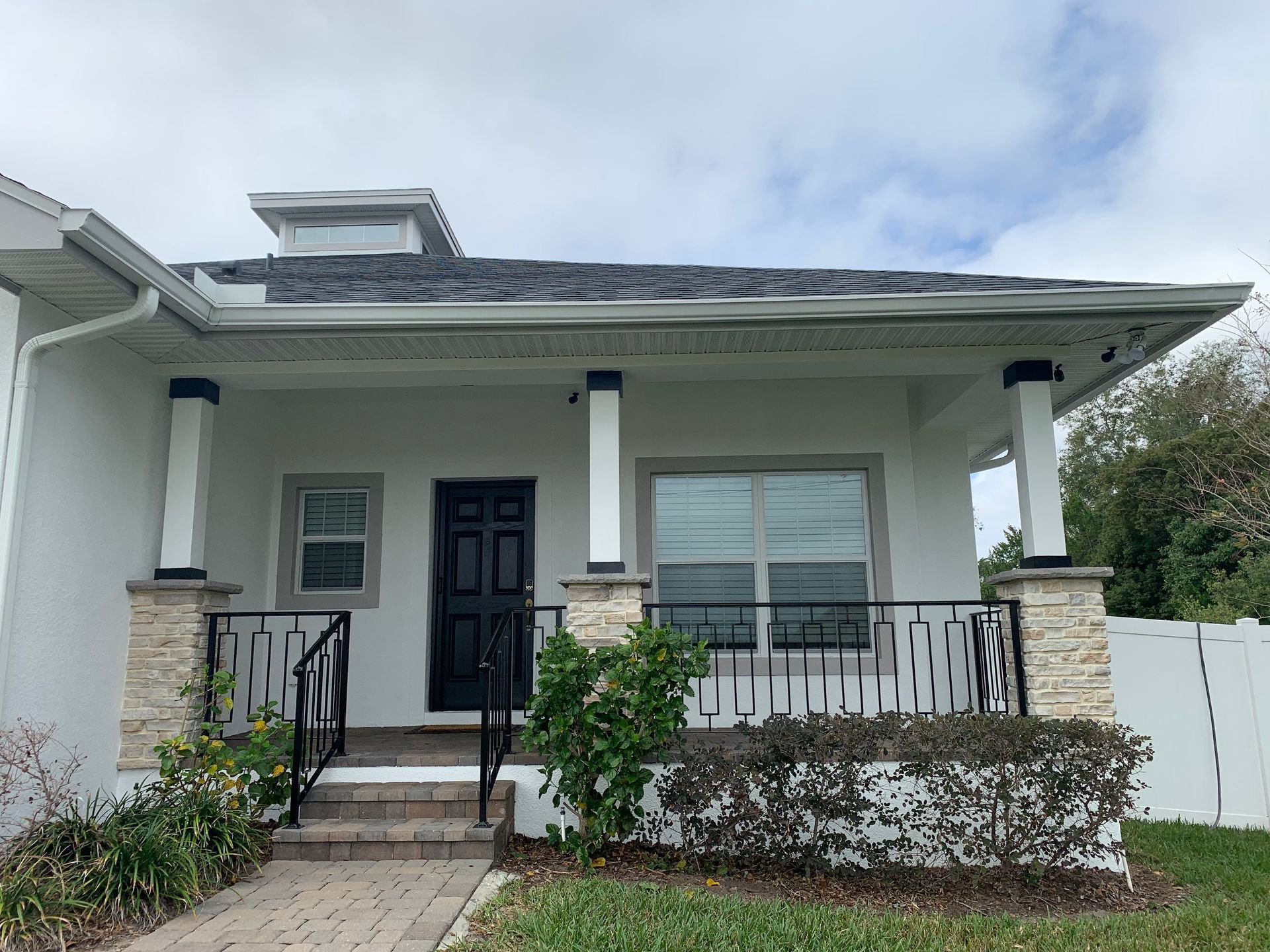 White house exterior with black door, pillars, and railing, featuring a brick pathway and green bushes.