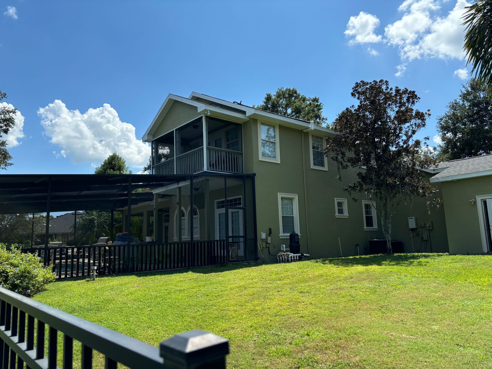 Two-story green house with screened balcony, covered patio, and green lawn under blue sky.