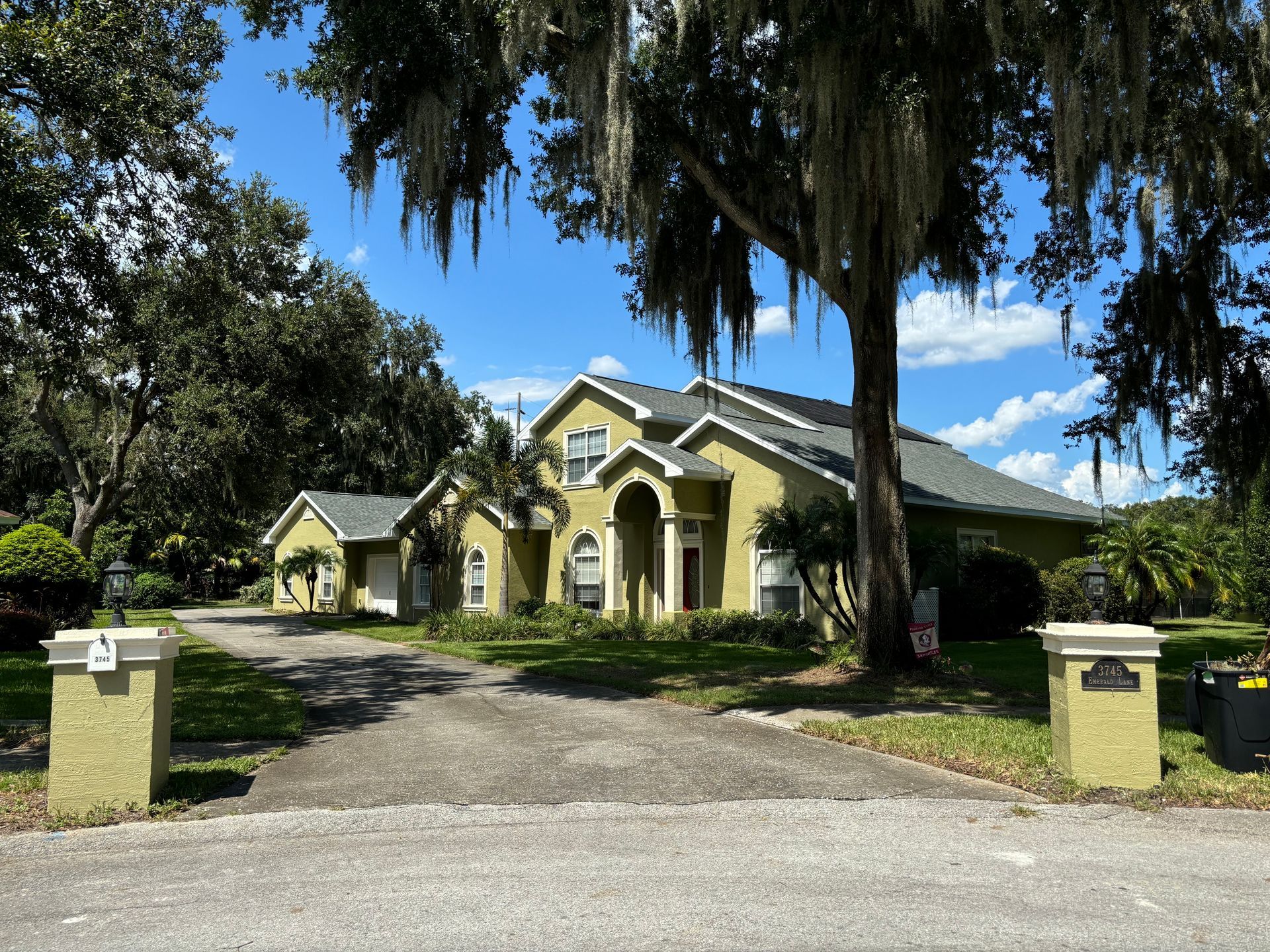 A two-story beige house with a long driveway framed by pillars, under a large tree.
