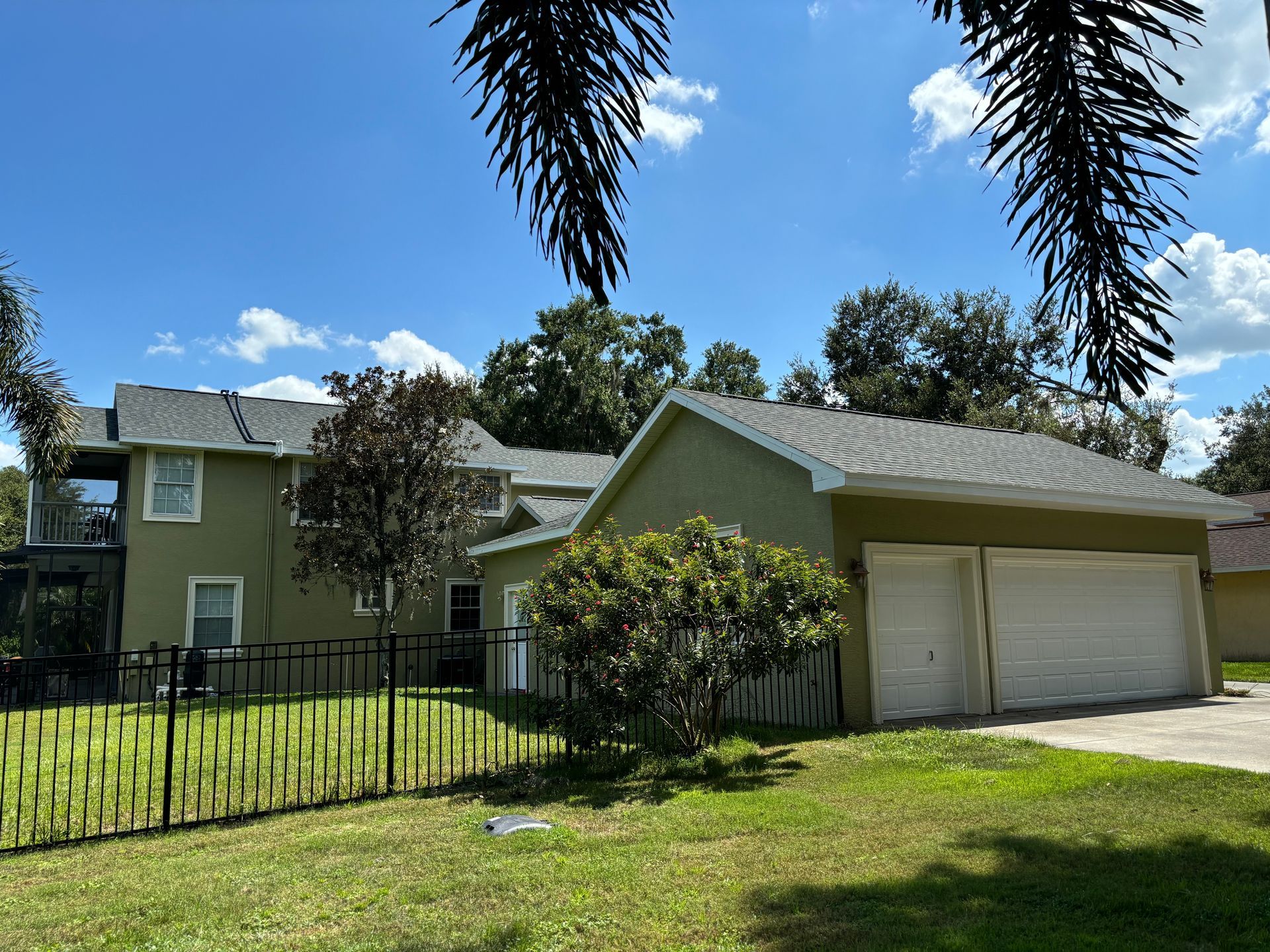 Green house with a two-car garage, black fence, and lush green lawn under a blue sky.