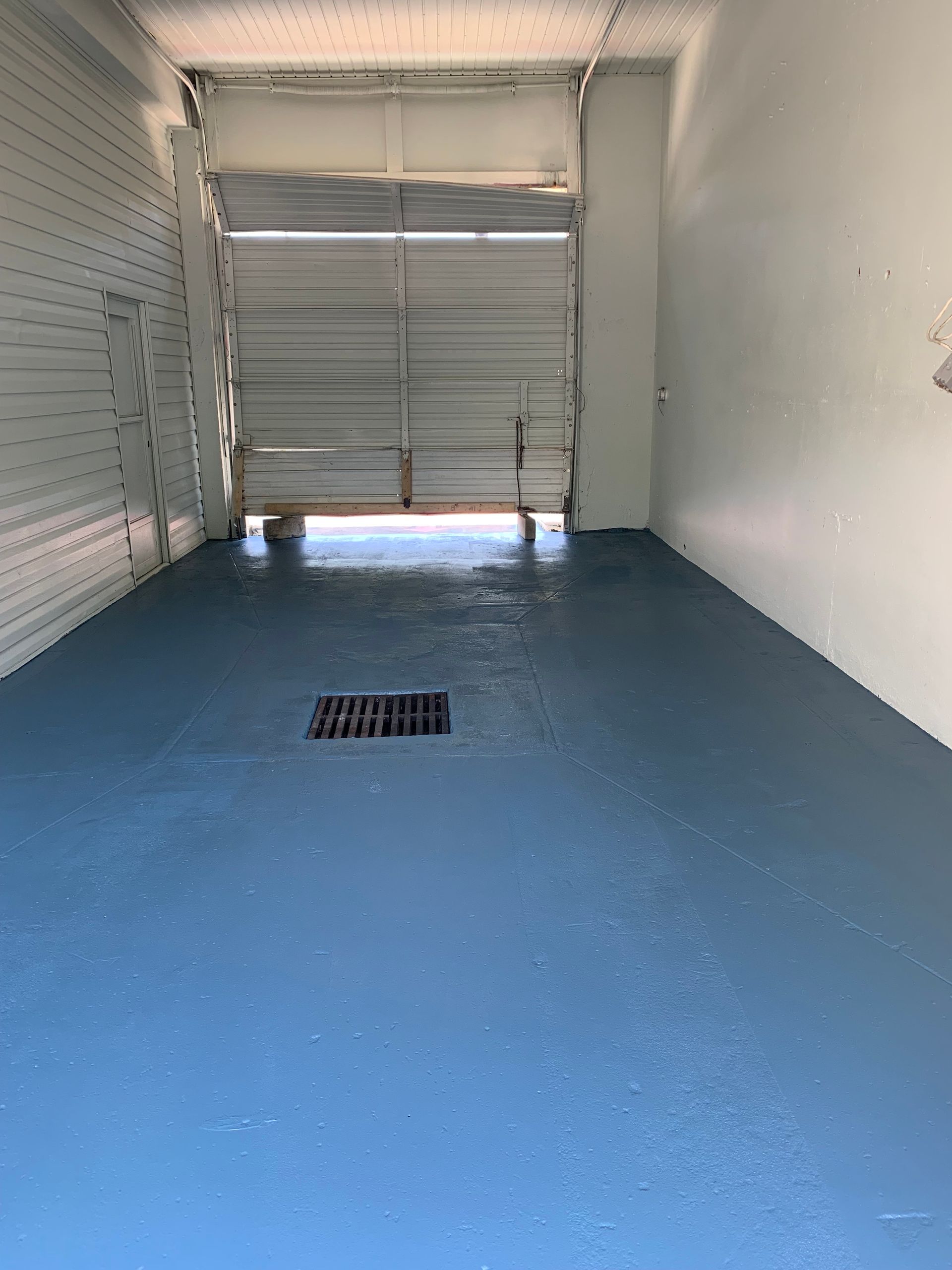 Blue-painted storage unit interior with open garage door, gray drain, and bright light.