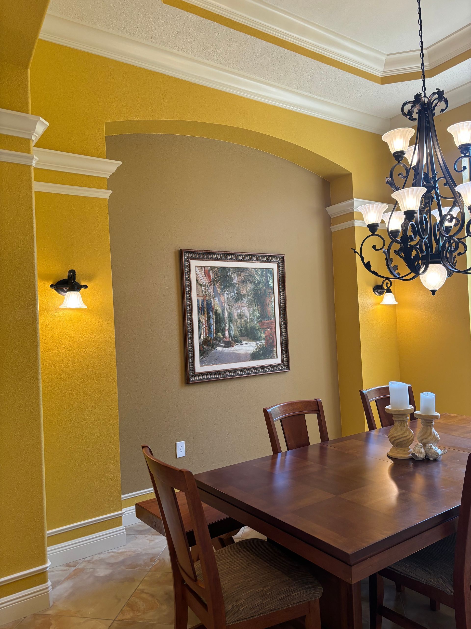 Dining room with yellow walls, a wood table, and ornate chandelier; artwork and sconces visible.