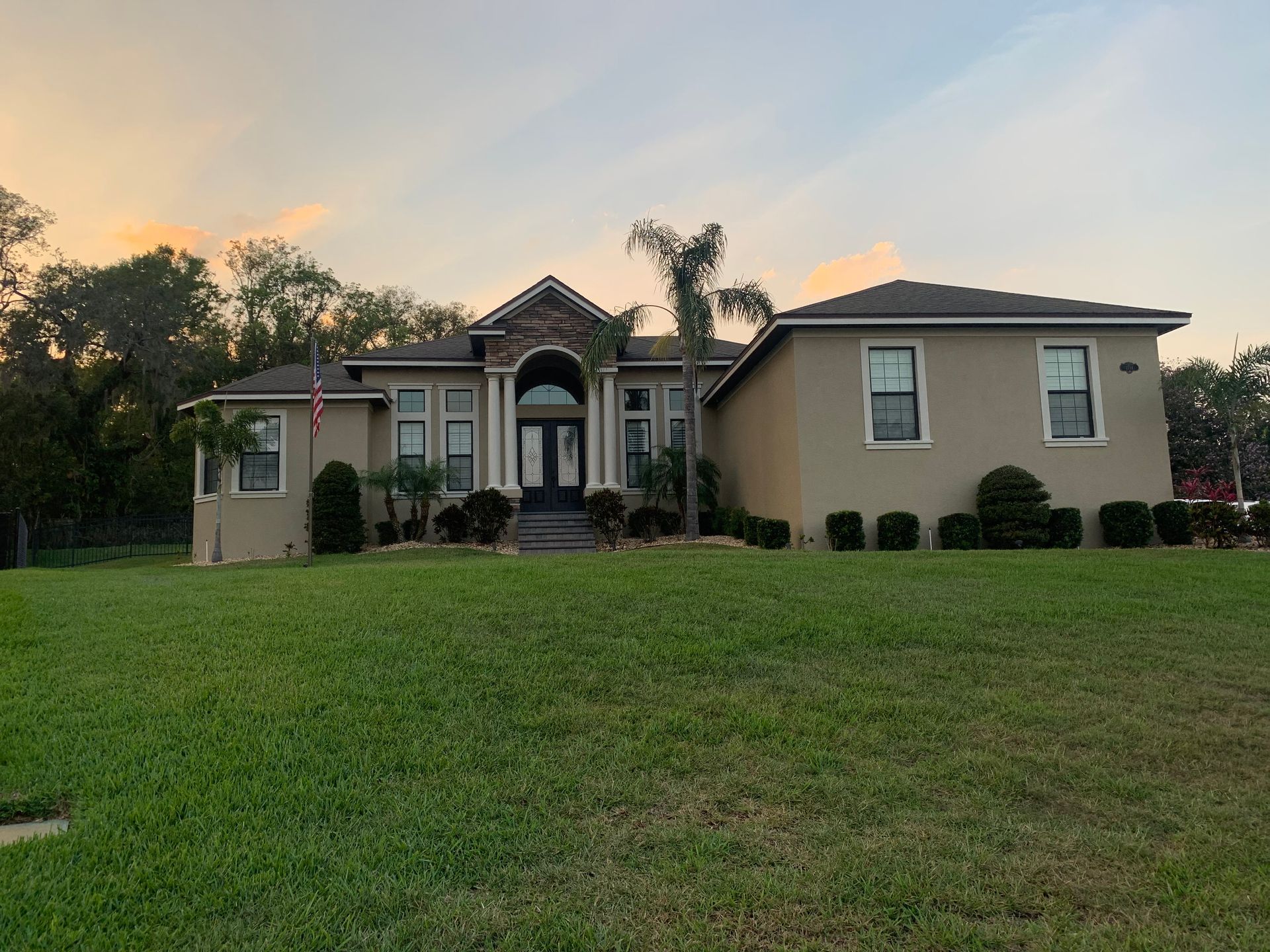 Tan house with black door, green lawn, and trees against a sunset sky.